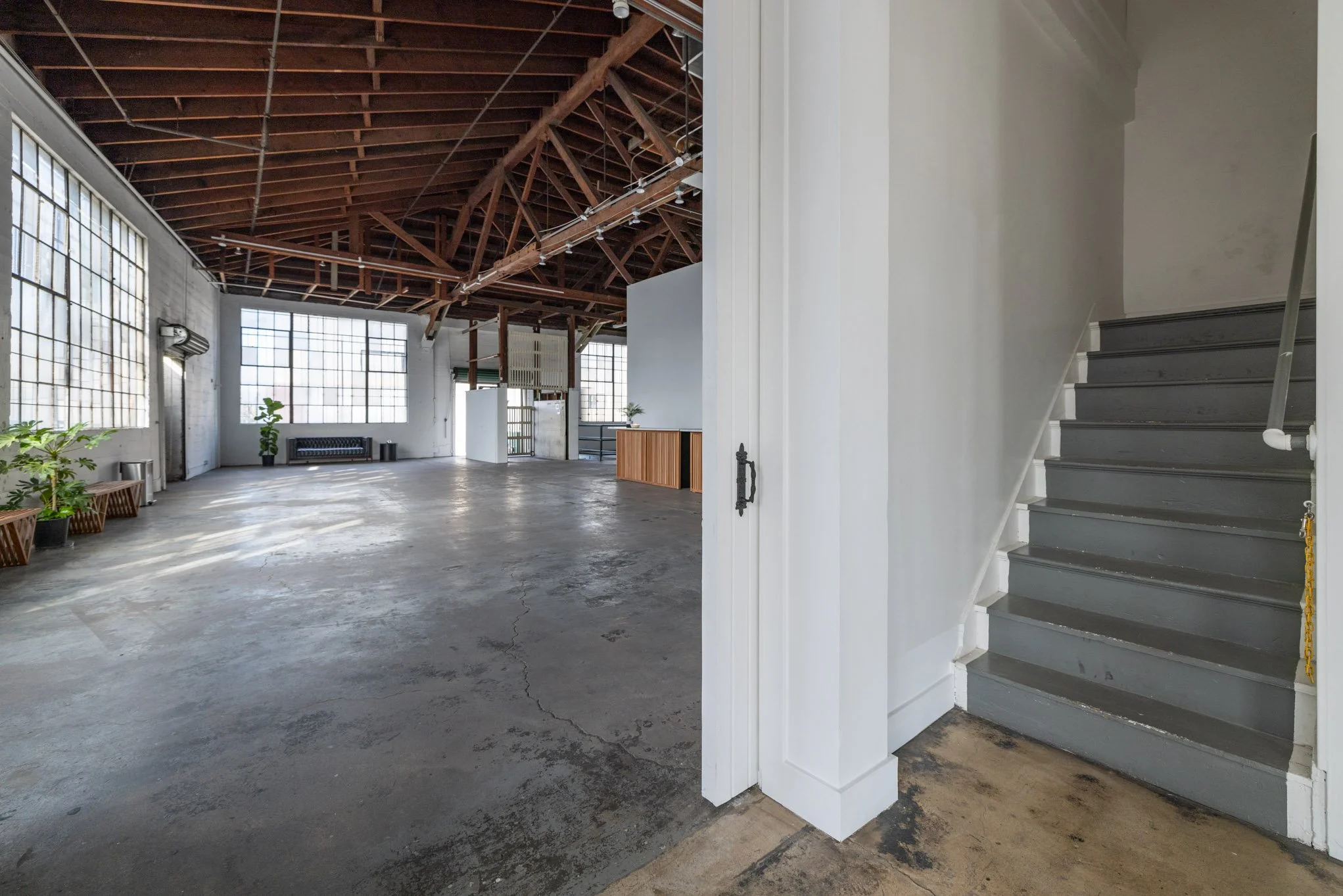 Interior view of a spacious, industrial-style loft with large windows, exposed wooden beams, and a concrete floor, with a small staircase on the right side.