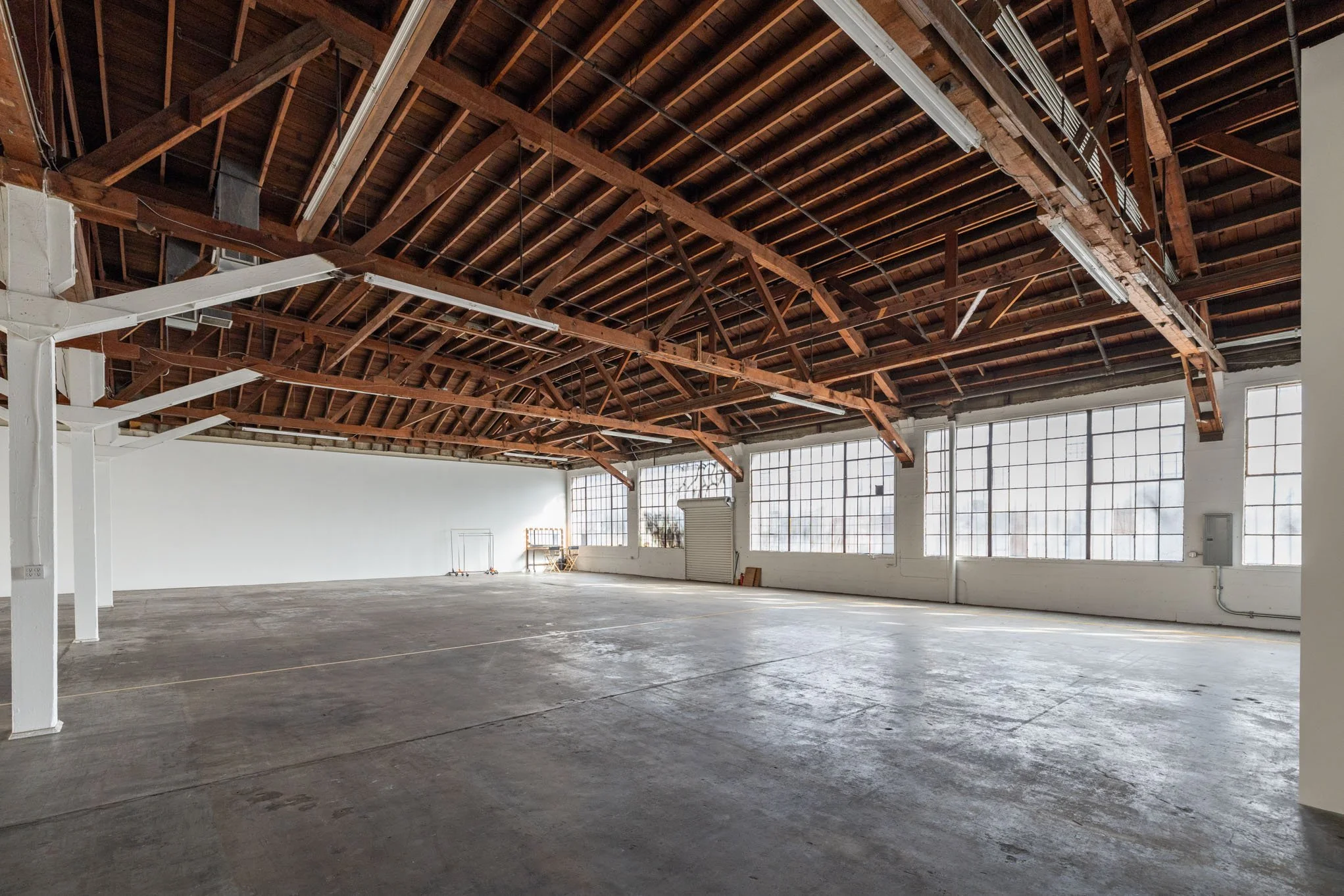 Empty industrial warehouse with large windows, exposed wooden ceiling beams, and concrete floor.