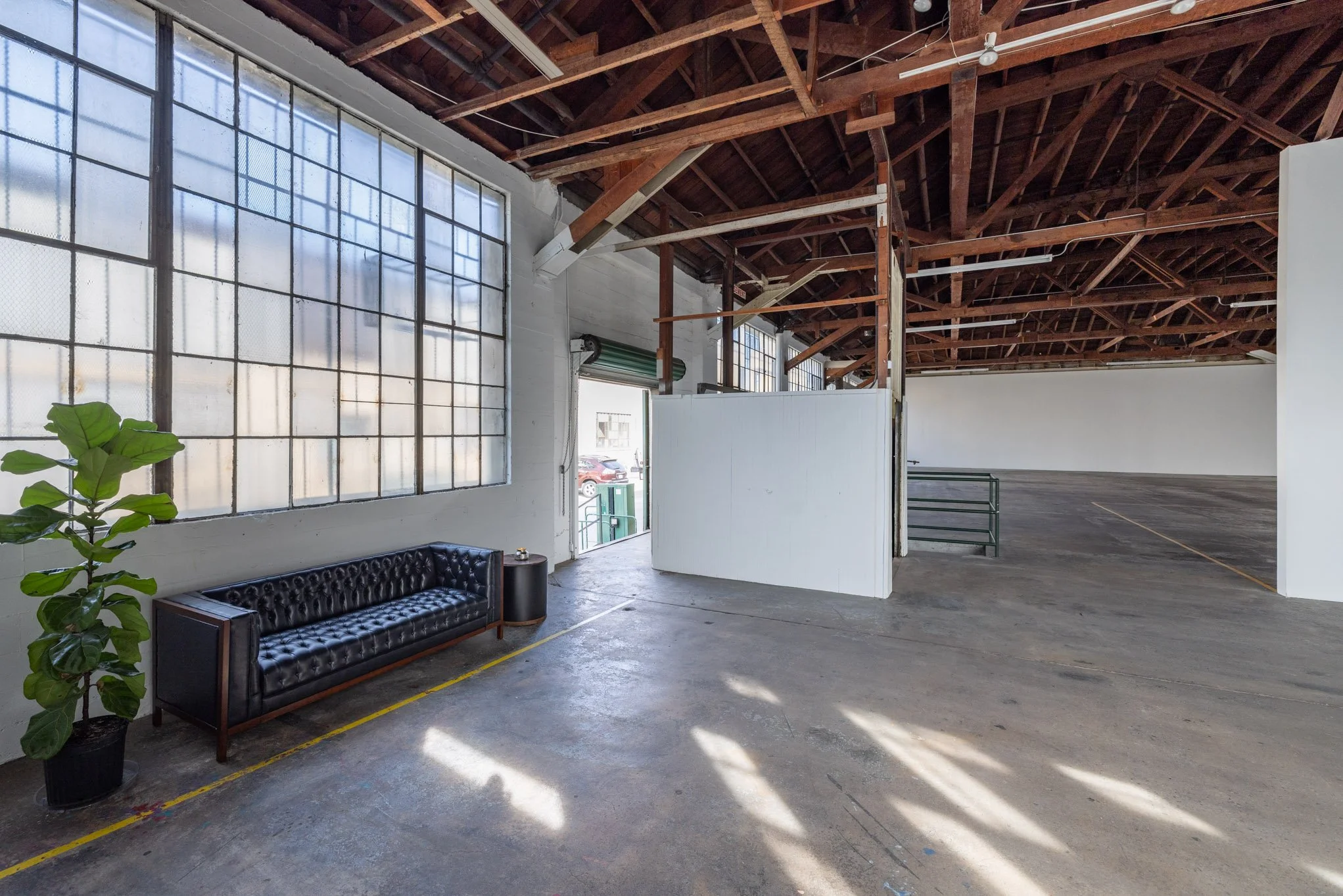 Empty industrial space with large windows, a black leather sofa, a small side table, a potted plant, and a white partition wall.