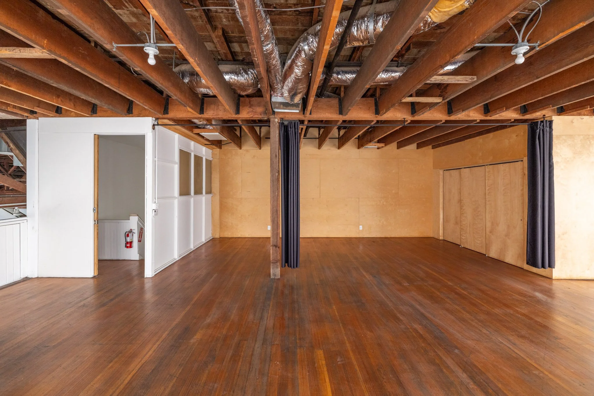 Empty open space with wooden floors, exposed ceiling beams, industrial ducts, and black curtains on the sides.