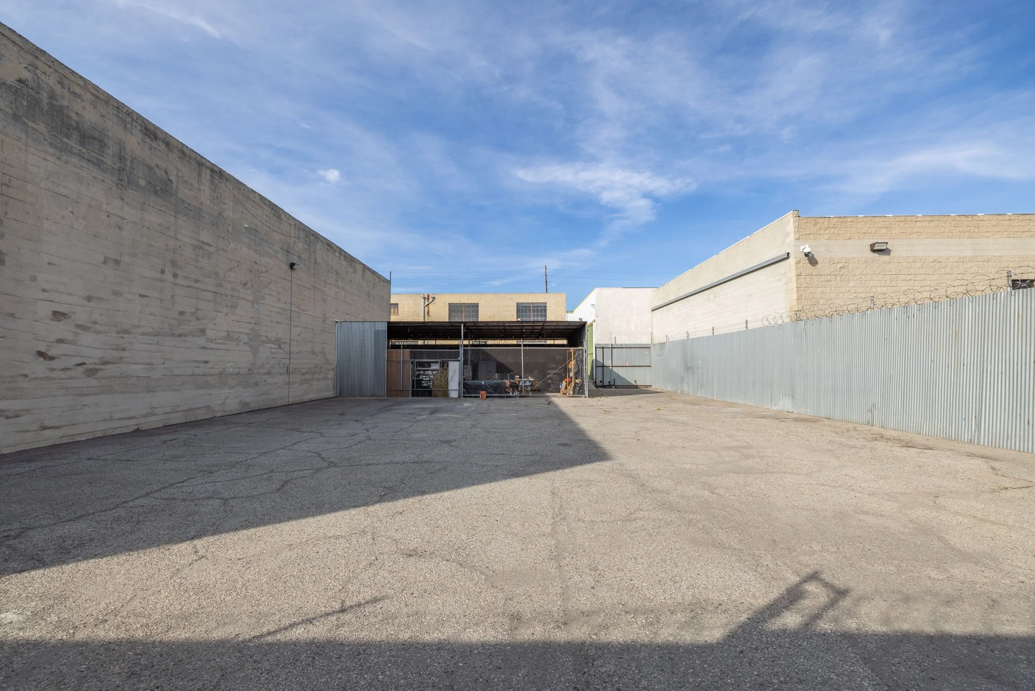 An empty outdoor lot with cracked asphalt, tall concrete walls on both sides, a chain-link fence with barbed wire surrounding a small structure, and cloudy blue sky.
