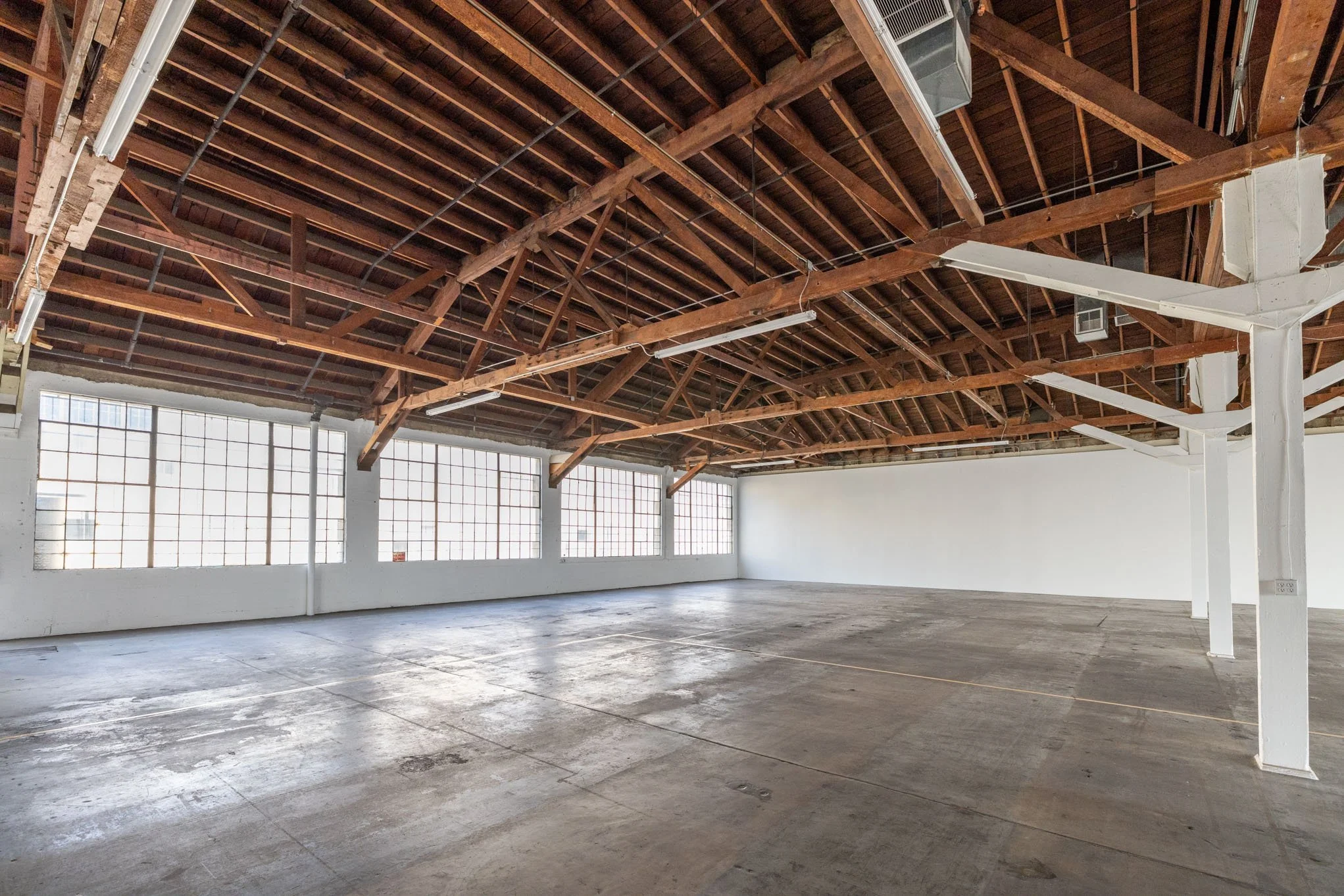 Empty industrial warehouse with large windows, wooden ceiling beams, and concrete floor.