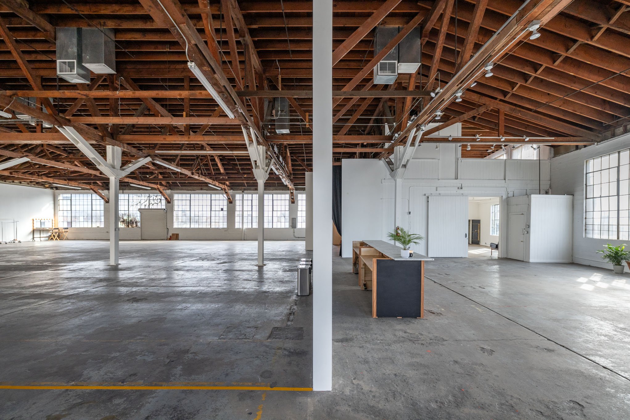 A split view of an empty, spacious industrial-style interior with large windows, high wooden ceilings, and concrete floors; the left side is open with minimal decor, and the right side has a small desk with potted plants and a door leading to another