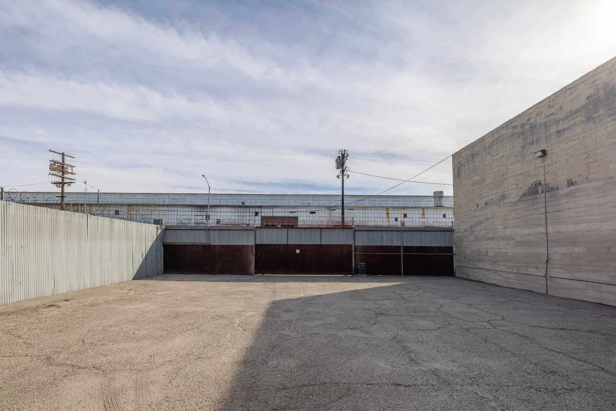 Empty outdoor parking lot enclosed by metal fencing and walls with utility poles and an industrial building in the background under a cloudy sky.