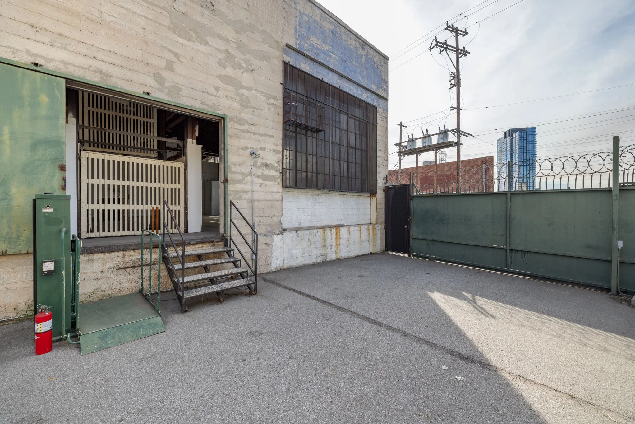 Back alley of an industrial building with metal stairs, a fire extinguisher, a small green platform, a green gate, and a large gray wall with barred window. Power lines and a city skyline are in the background.