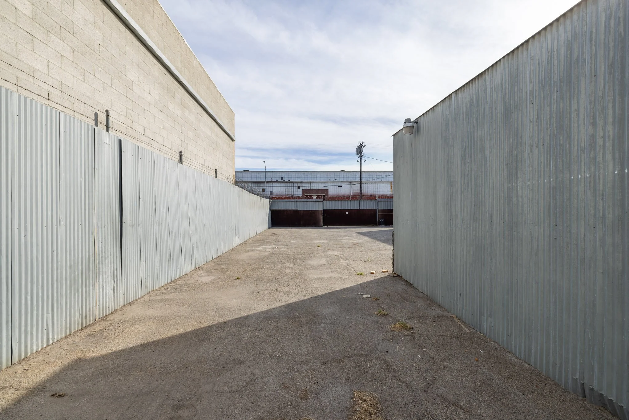 An empty alleyway enclosed by tall metal fences on both sides, with a building in the background and a cloudy sky overhead.