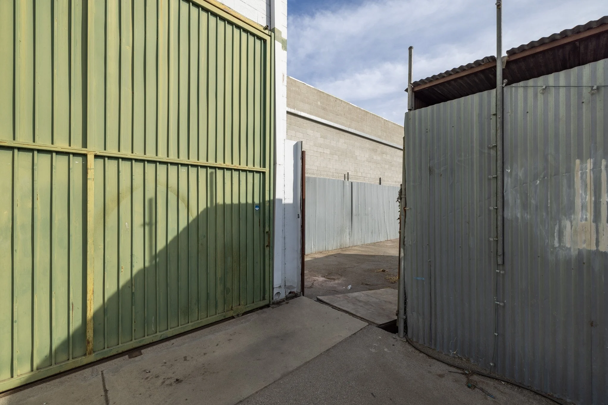 Open metal gate with green panels on the left and a curved gray panel on the right, leading into an alley with concrete and asphalt surfaces, surrounded by a fence and buildings under a partly cloudy sky.