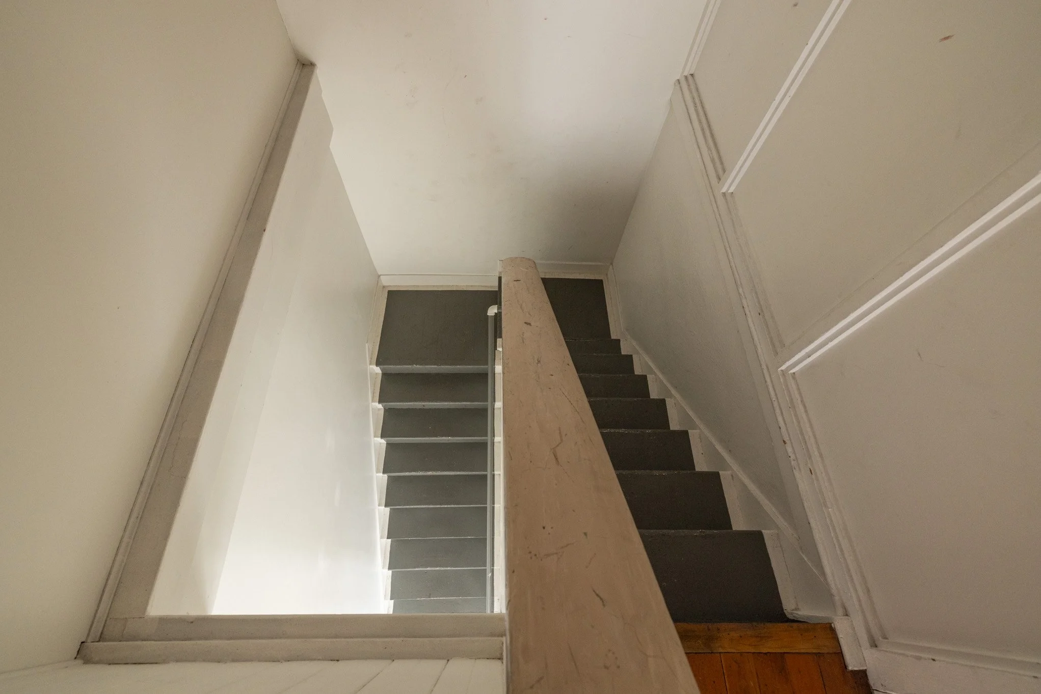 Indoor staircase viewed from the top, with dark gray steps, white walls, and a beige wooden railing.