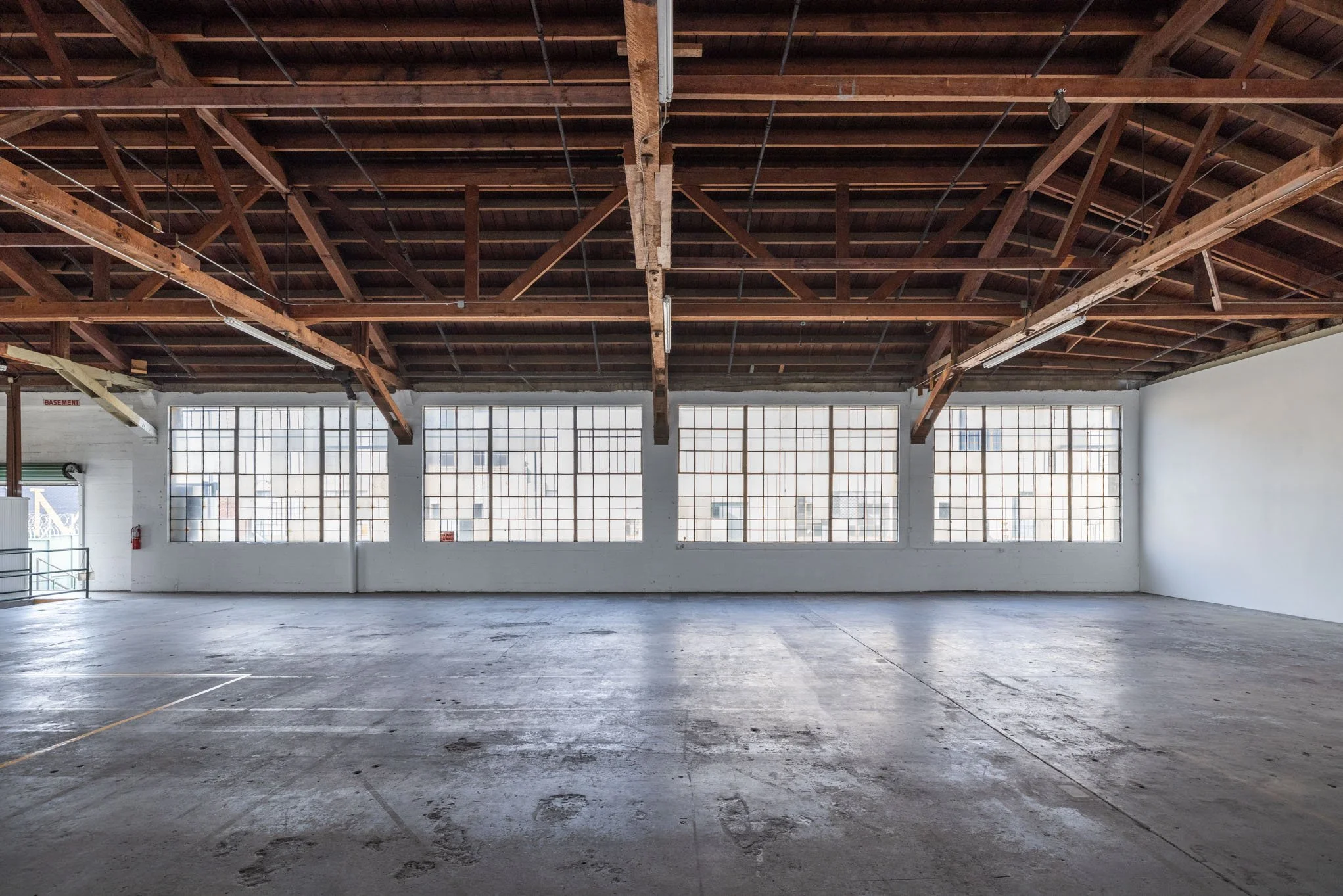 Empty industrial or warehouse space with large grid windows, concrete floor, exposed wooden ceiling beams, and a small staircase on the side.