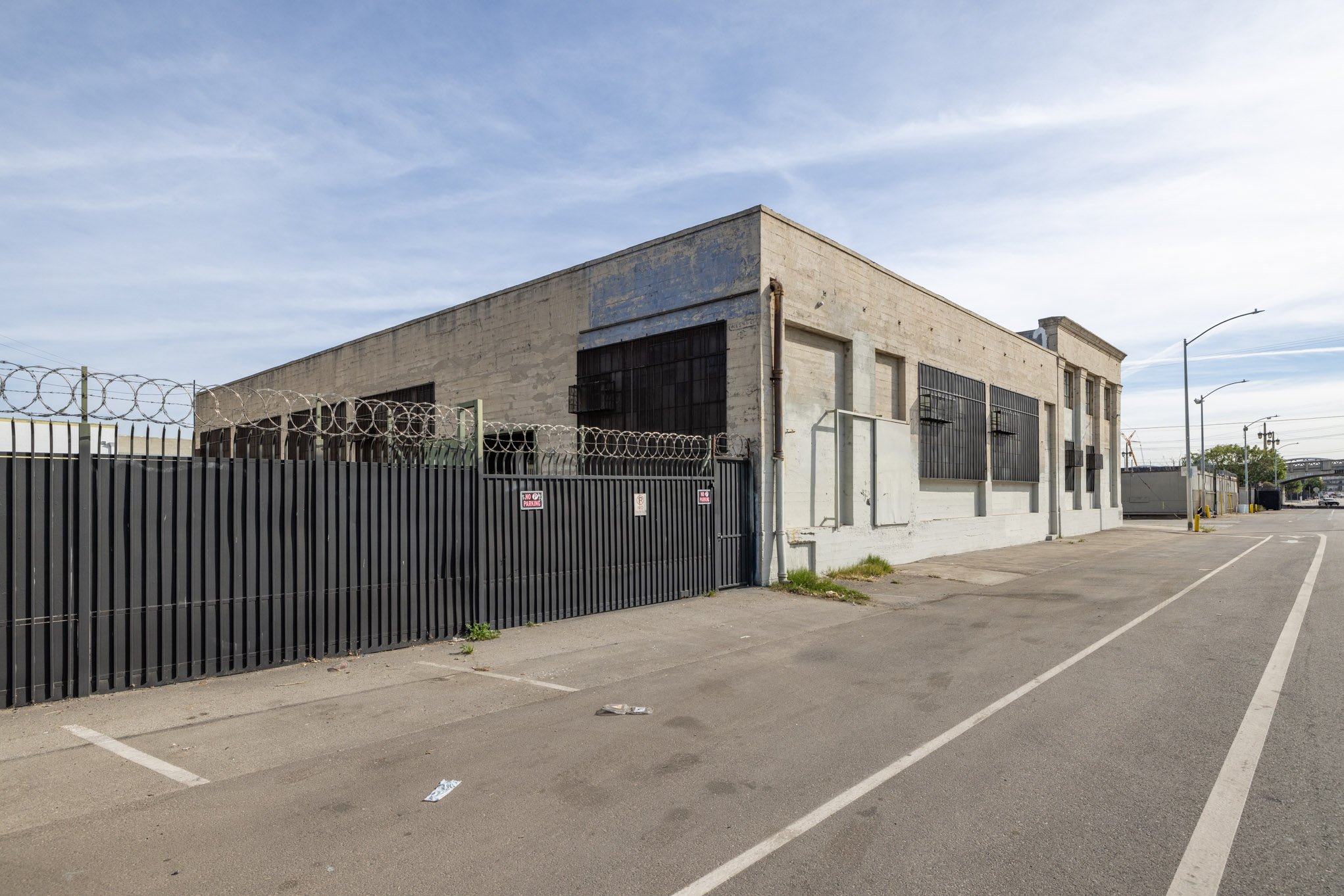 An empty parking lot next to a beige industrial building with barred windows, surrounded by a black fence topped with barbed wire, under a partly cloudy sky.