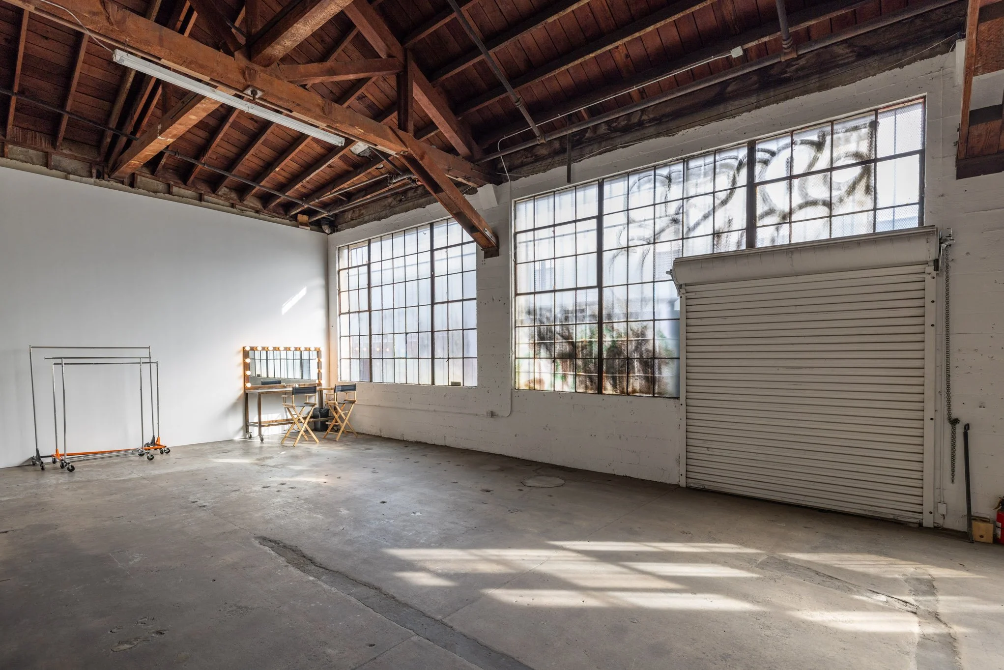 Empty industrial-style room with large windows, concrete floor, exposed wooden ceiling beams, and minimal furniture including a makeup mirror with lights, two chairs, and a rolling clothes rack.