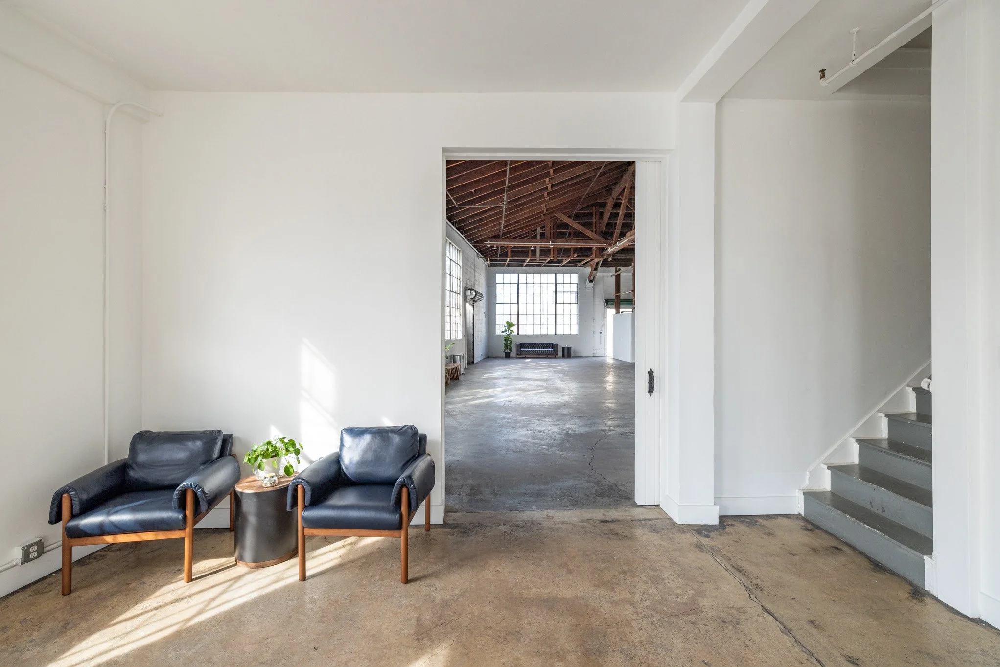 Interior of a minimalist, industrial-style living space with two black leather chairs and a small round table with a plant, leading into a larger room with exposed wooden ceiling beams and large windows.