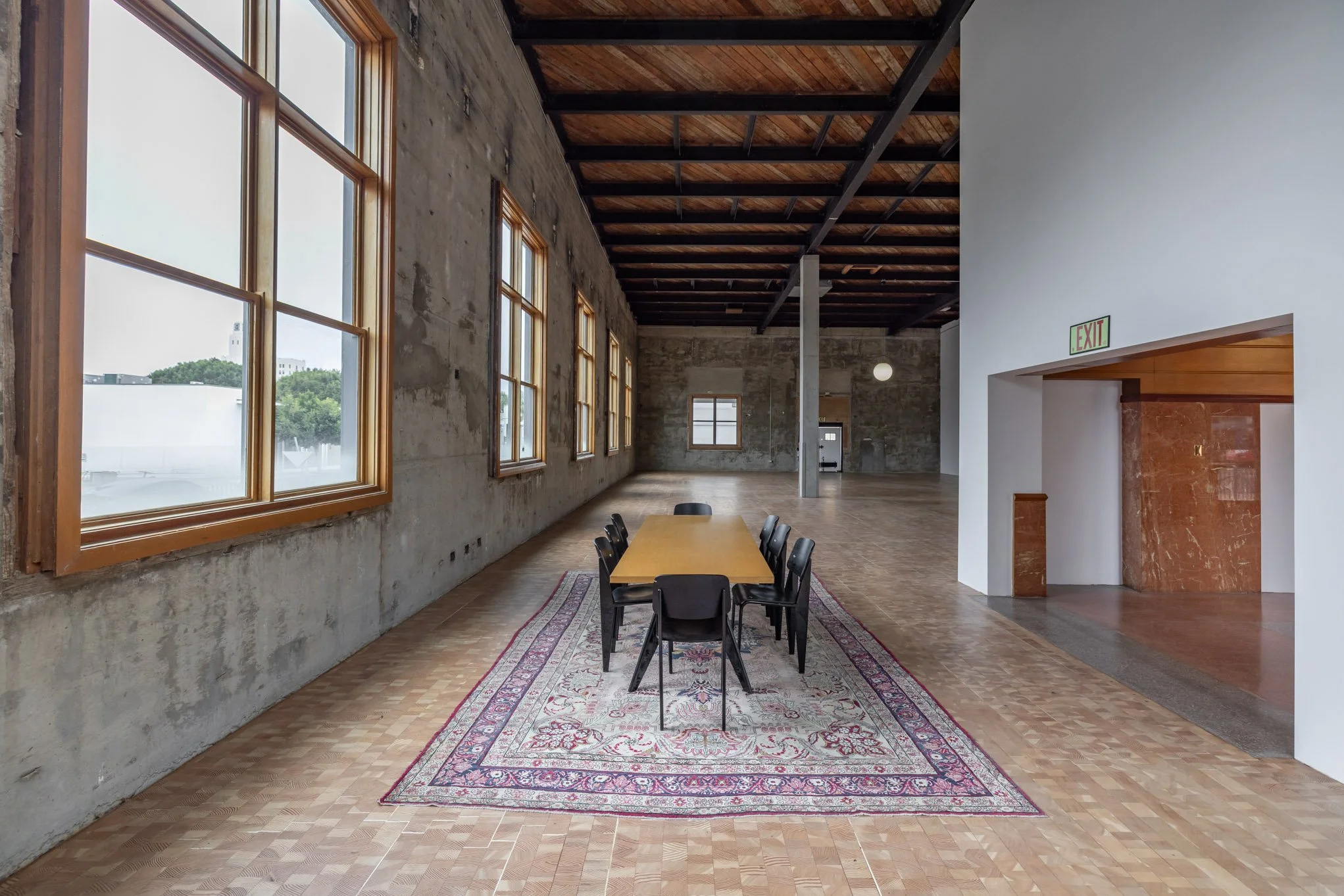 Empty conference room with wooden flooring, large windows, exposed brick walls, and a long table with eight chairs on an ornate rug.