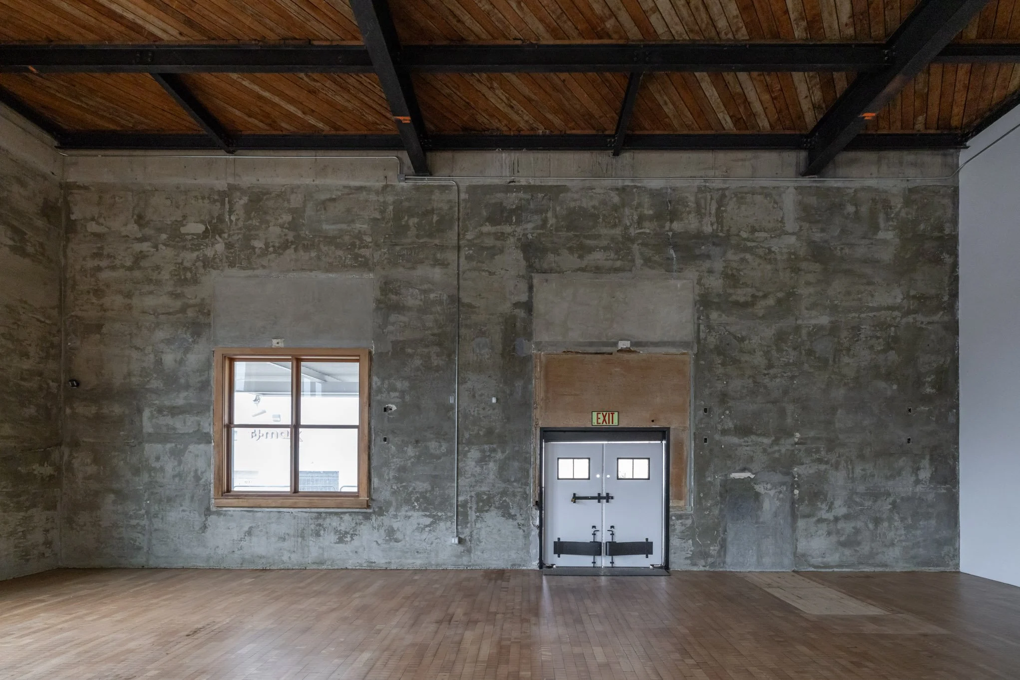 Empty industrial-style room with a concrete wall, a window with a wooden frame, a metal exit door, and a wooden floor.
