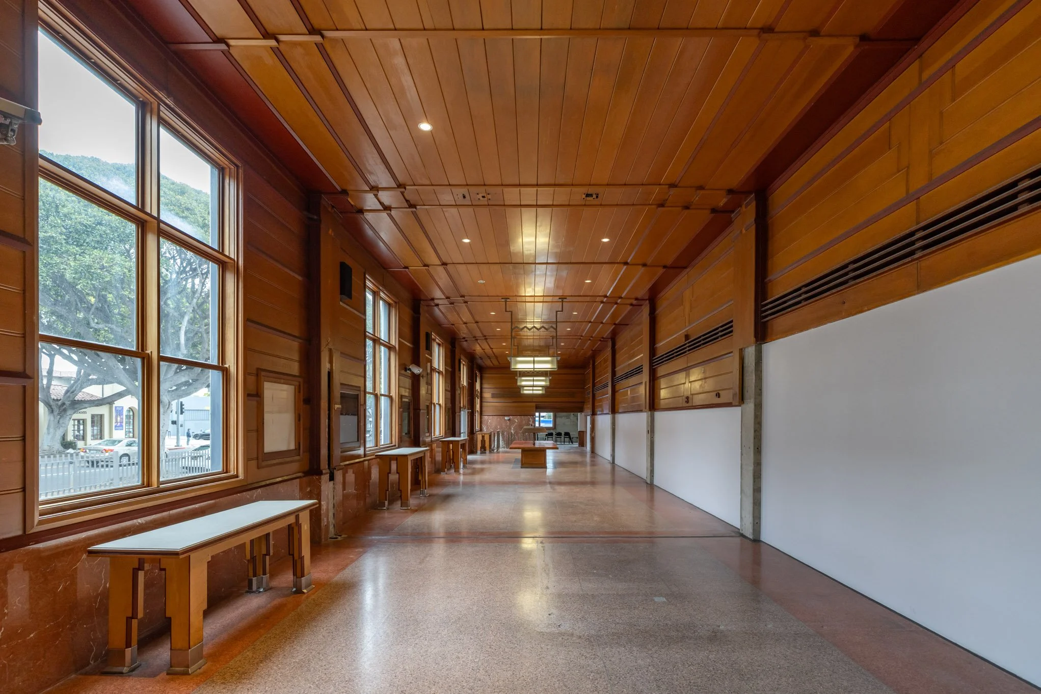 Empty hallway with wooden paneling on walls and ceiling, with benches along the windows and hanging ceiling lights.