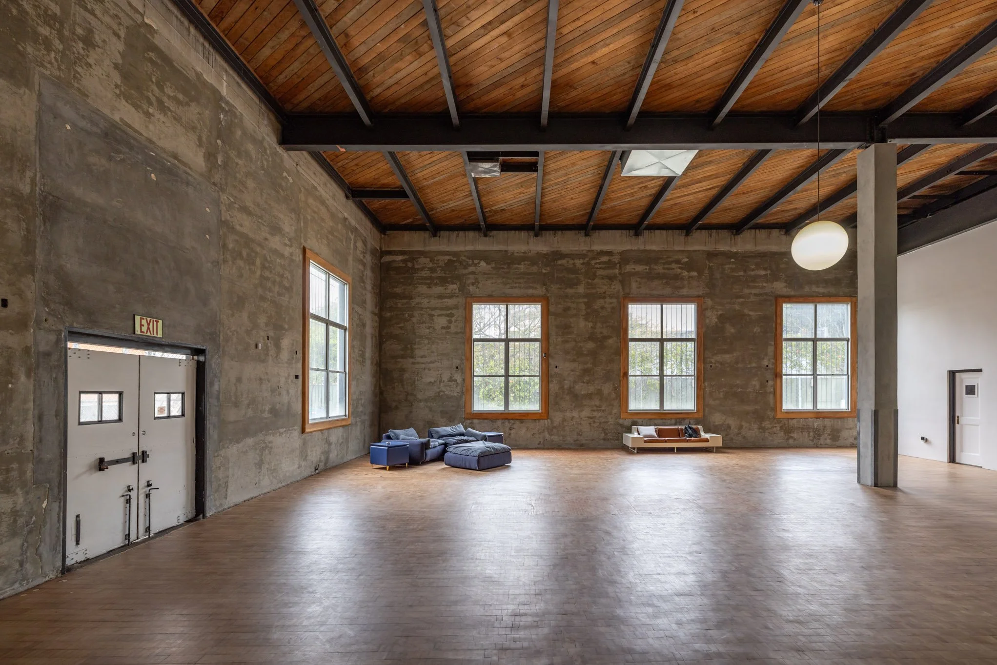 An empty industrial-style room with concrete walls, a wooden ceiling, large windows, and minimal furniture including a blue sectional sofa and a lounge chair.