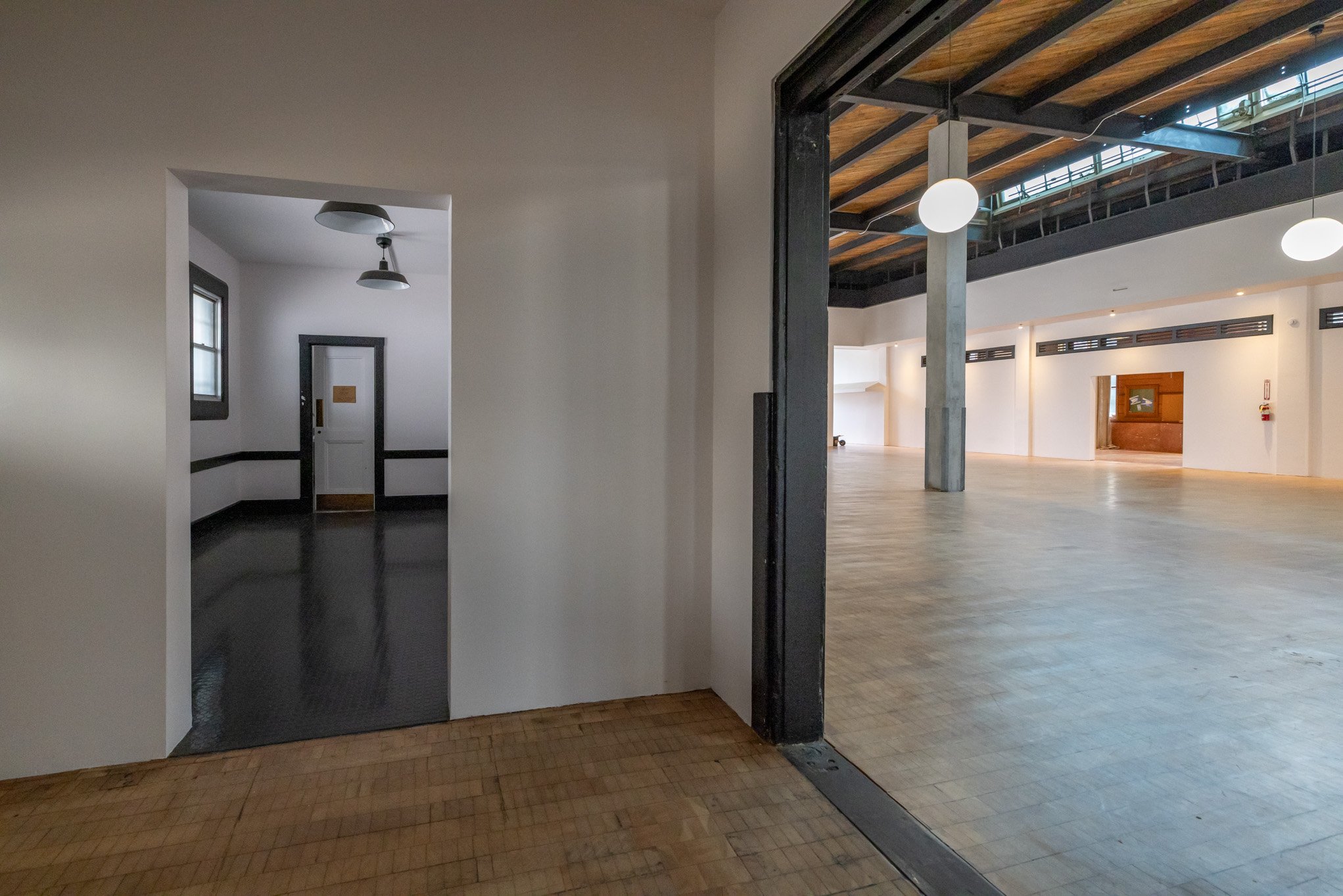 Interior of an empty modern building with white walls, wooden and black floors, high ceilings, and large open spaces illuminated by spherical hanging lights.