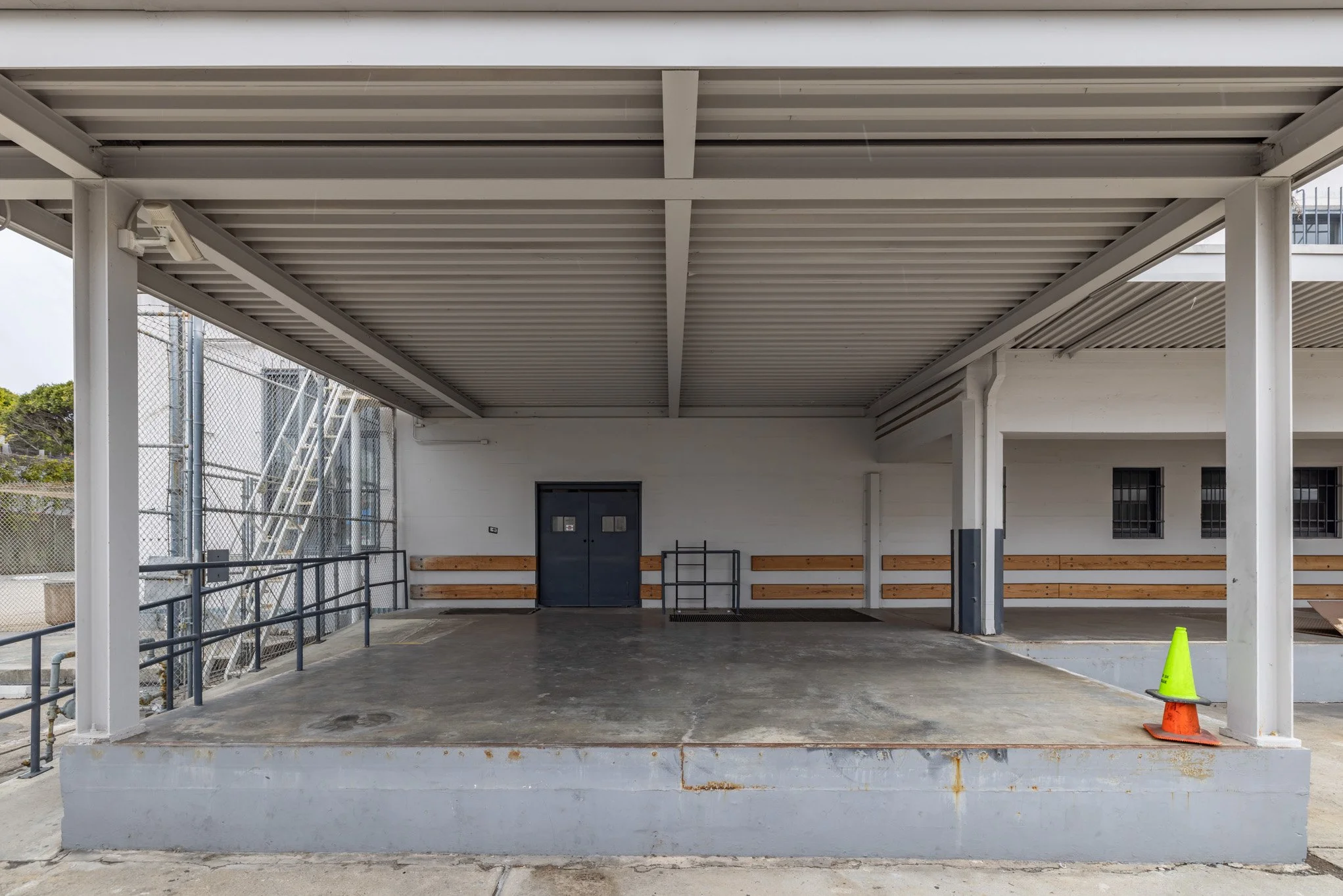 Empty outdoor shelter with a concrete floor, metal railing, benches, a door, and construction cones.