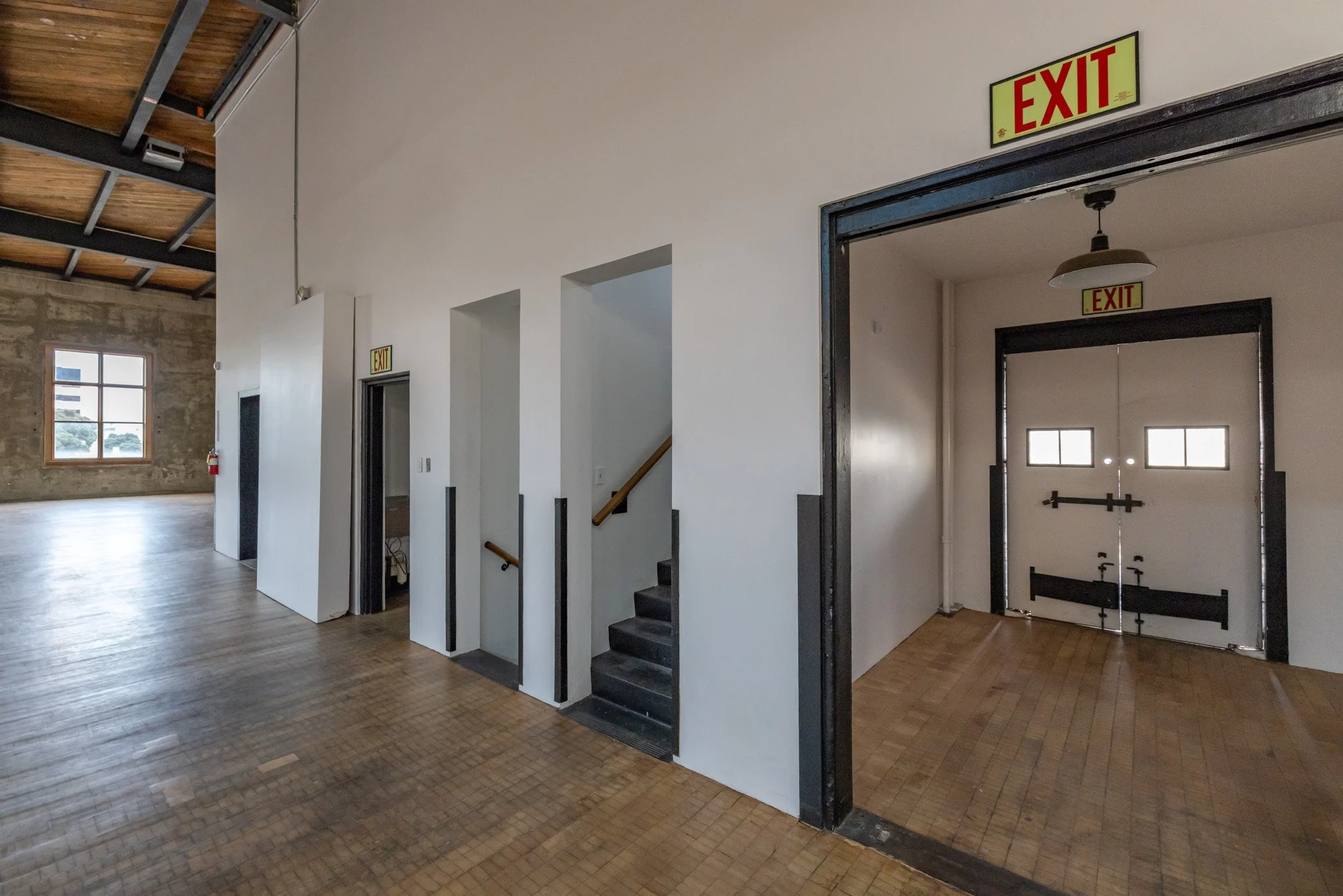 Interior of a building with multiple exit doors, a staircase, and a large window, featuring exposed ceiling beams and wooden flooring.