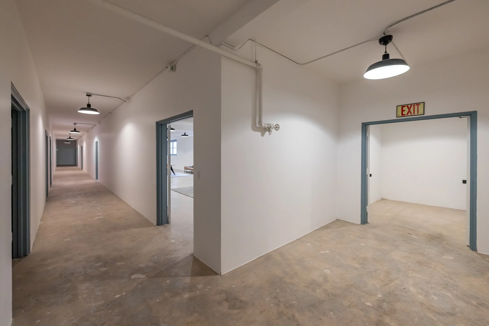 Empty hallway with multiple doors, some open, white walls, unfinished flooring, ceiling lights, and an exit sign above an open doorway.