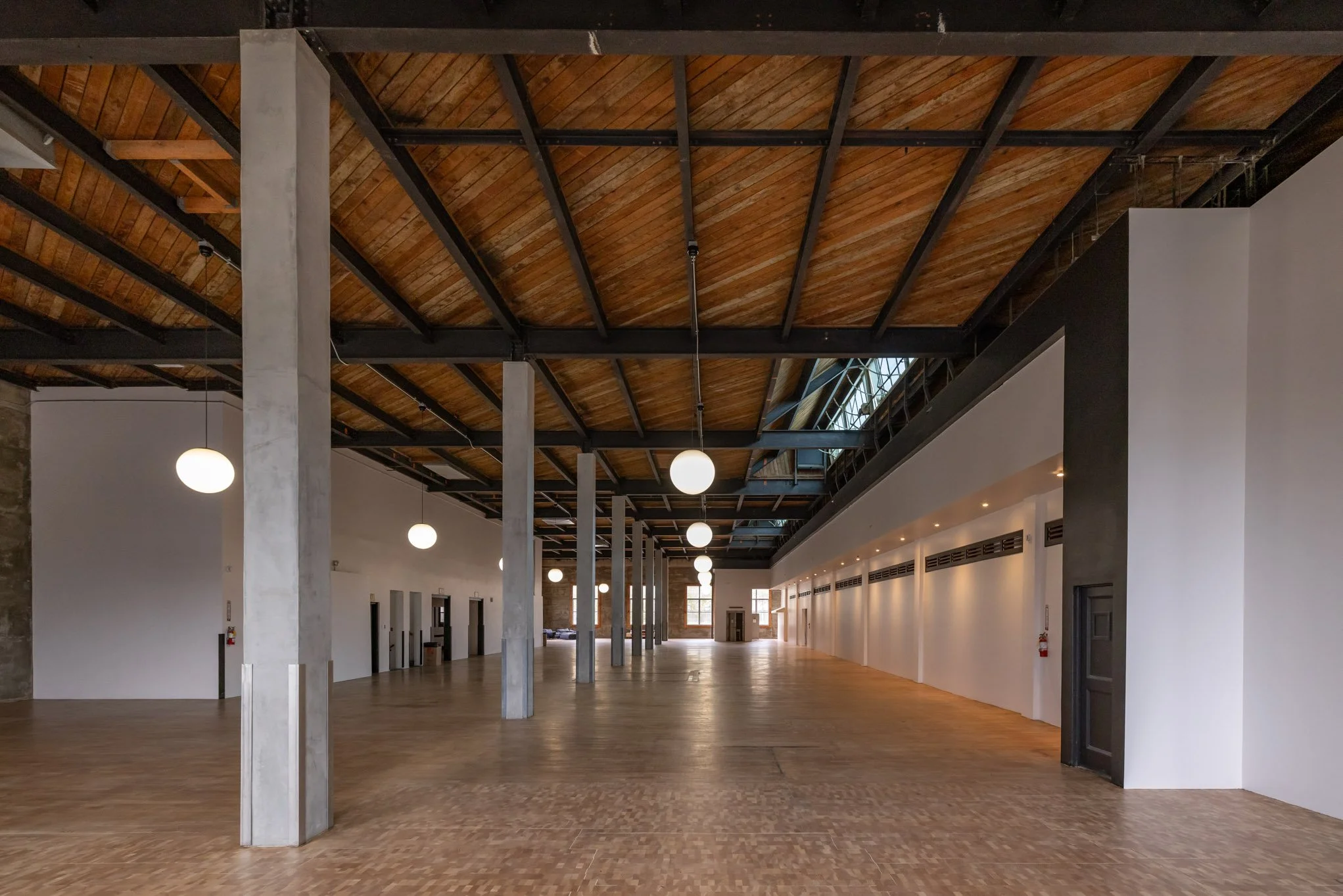Spacious interior with wooden flooring, white walls, black metal support beams, and hanging round ceiling lights, featuring large windows at the far end.