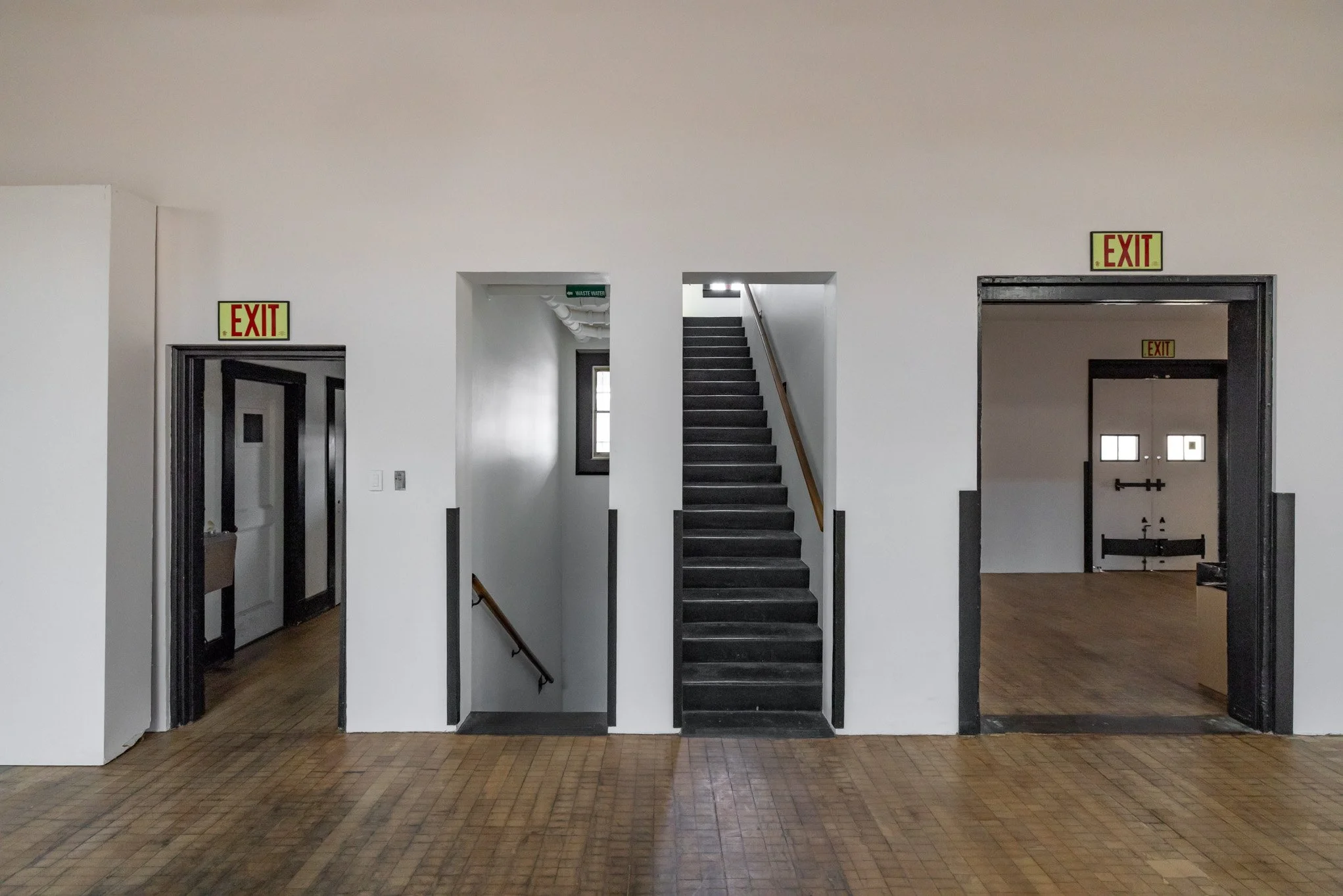 Interior view of a building with white walls, wooden floor, three doorways with 'EXIT' signs, and stairs in the middle leading upward.