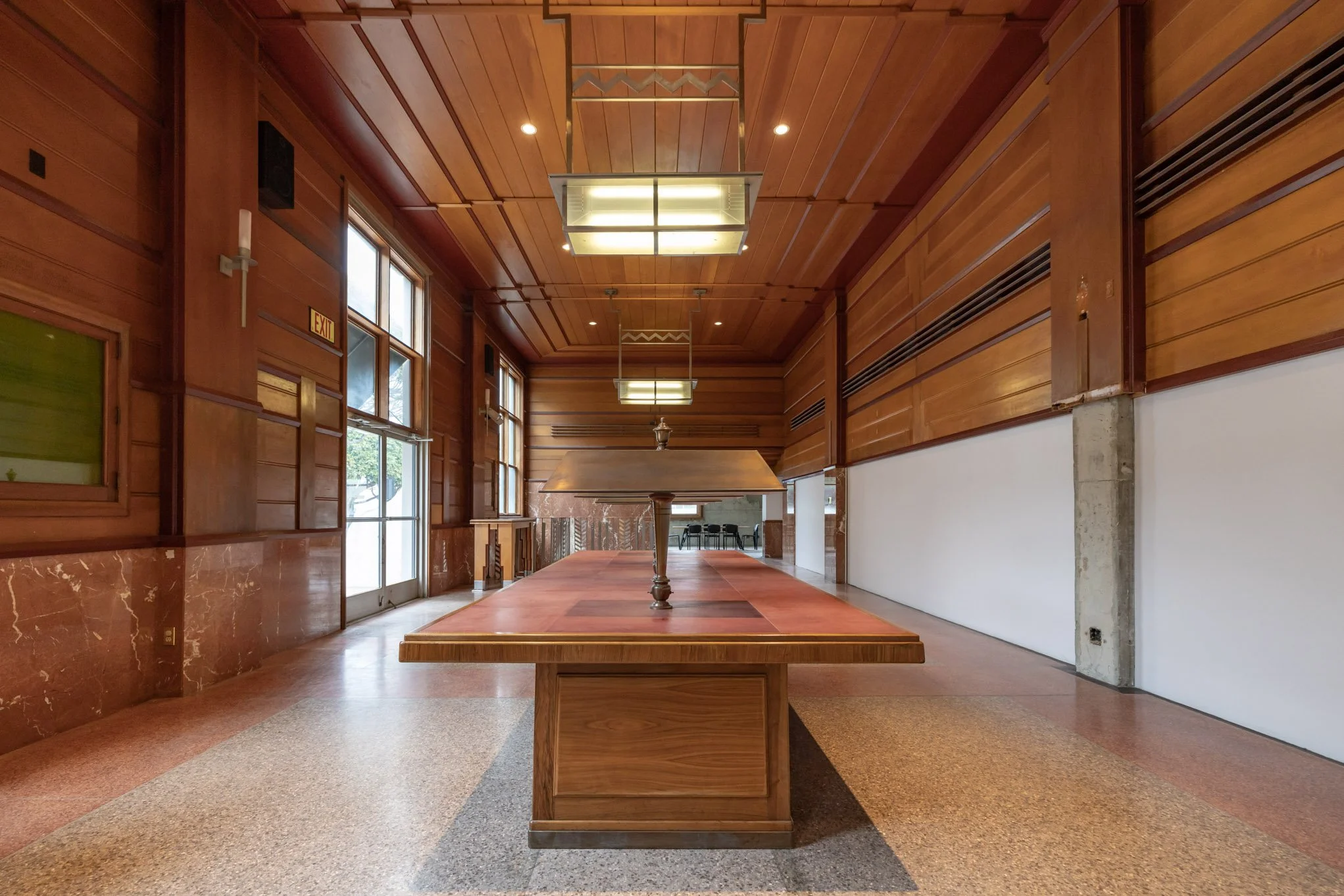 Empty indoor hall with wooden walls and ceiling, a large wooden table with a lamp on it, windows on the left side, and chairs in the background.