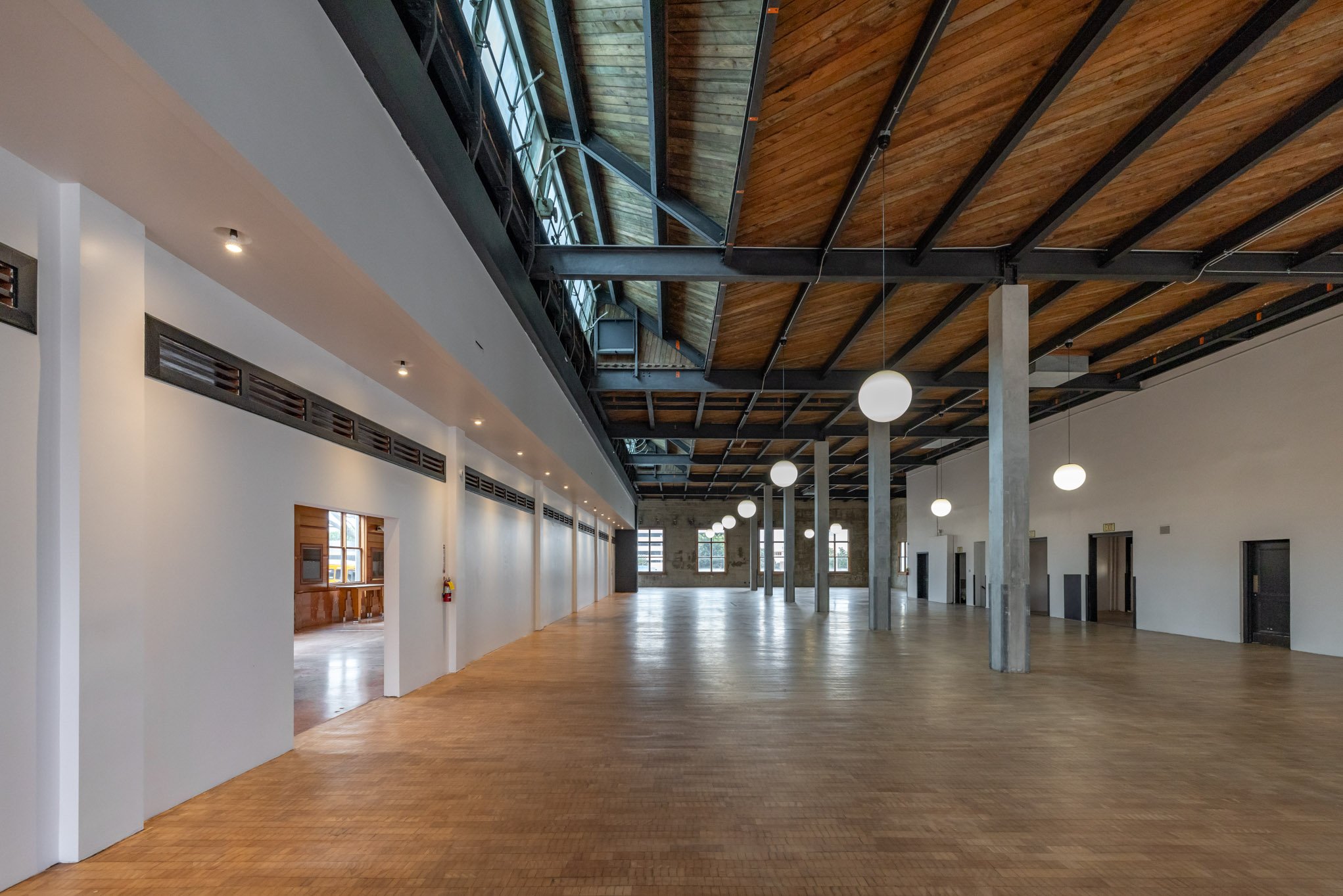 Empty spacious indoor hall with wooden floor, white walls, exposed ceiling with wooden panels, large windows, hanging spherical lights, and concrete support columns.