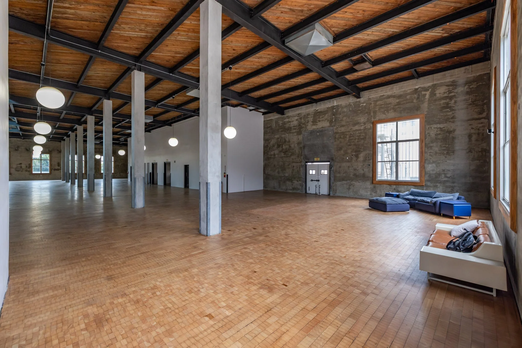 Spacious empty industrial-style room with wooden flooring, concrete walls, large windows, and hanging globe lights, with a few couches near the windows.