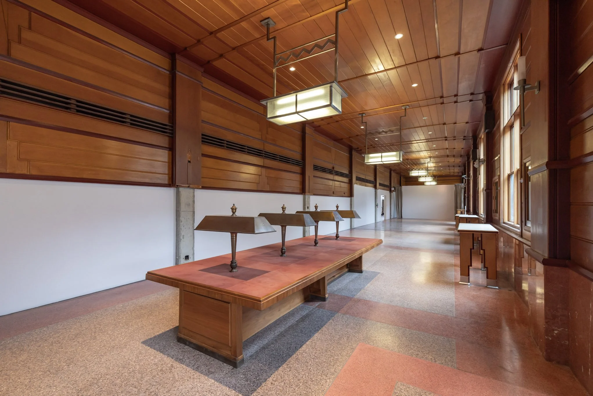 An empty, well-lit room with wooden walls, ceiling, and furniture, featuring a long wooden table with three brass lamps and large windows along one wall.