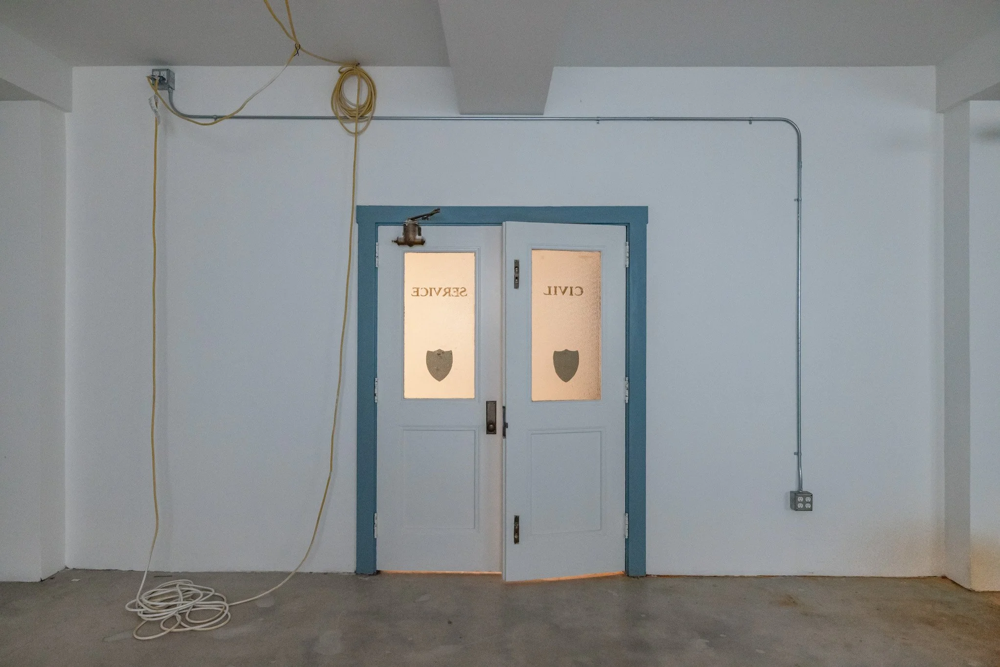 White interior wall with blue double doors labeled 'SERVIC' and 'CIVIL,' featuring shield emblems, with exposed electrical wiring hanging from the ceiling and a power outlet on the wall to the right.