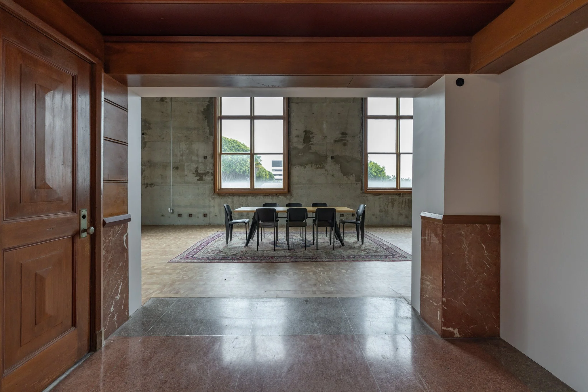 A room with a concrete wall, large windows, a wooden table, and chairs, with a patterned rug beneath the table.