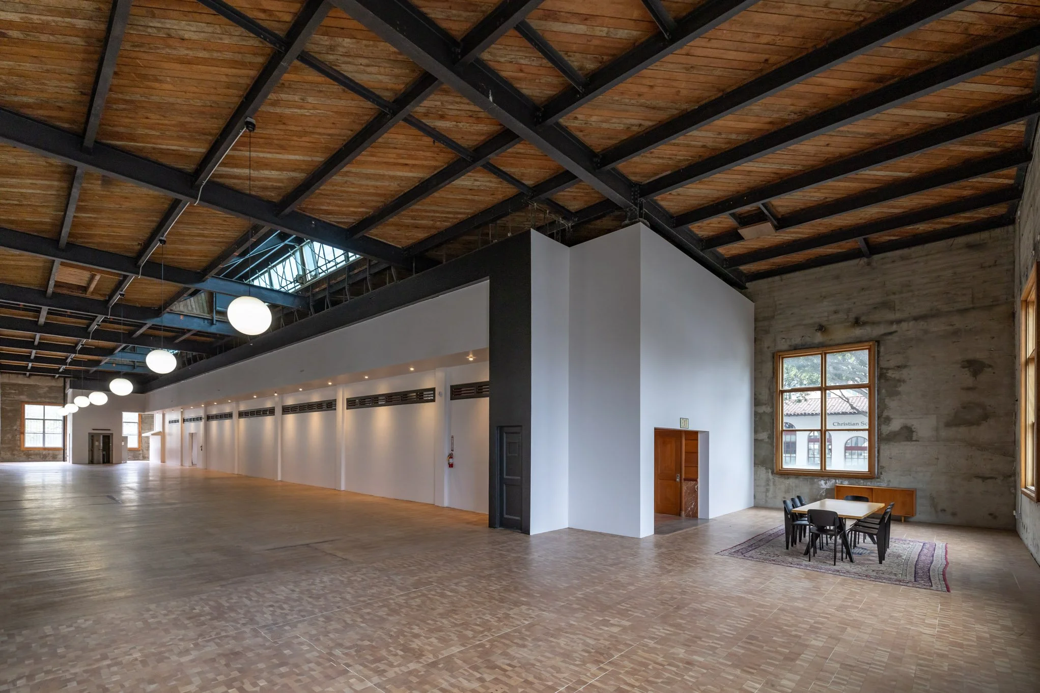 Spacious industrial-style interior with high wooden ceiling, black metal beams, large windows, and a small table with chairs near the wall.