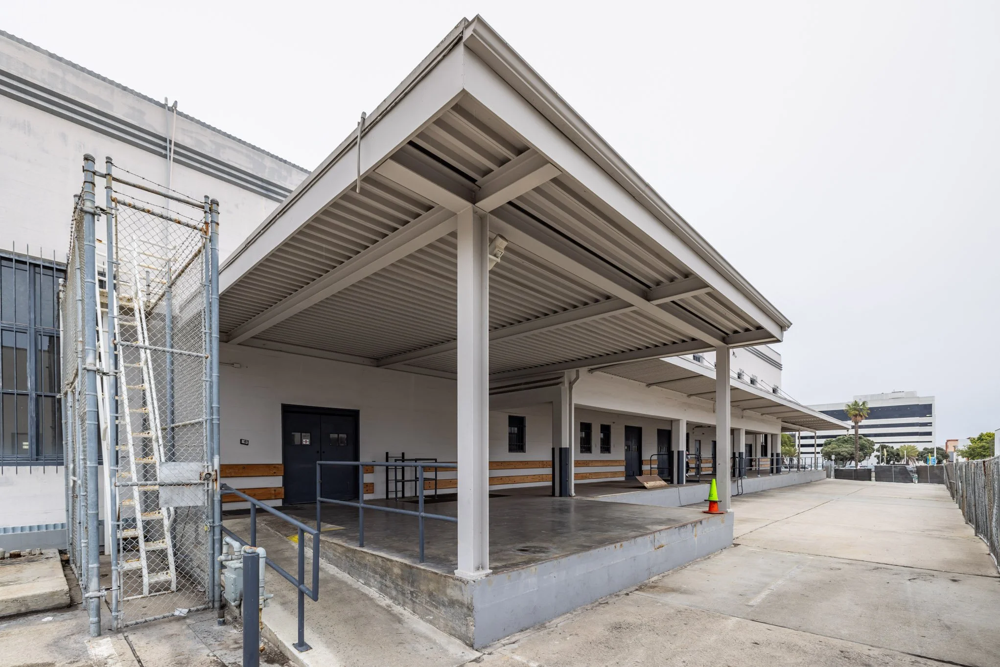 Empty loading dock area with overhead roof, black doors, safety railings, and an orange safety cone, adjacent to a fenced walkway.