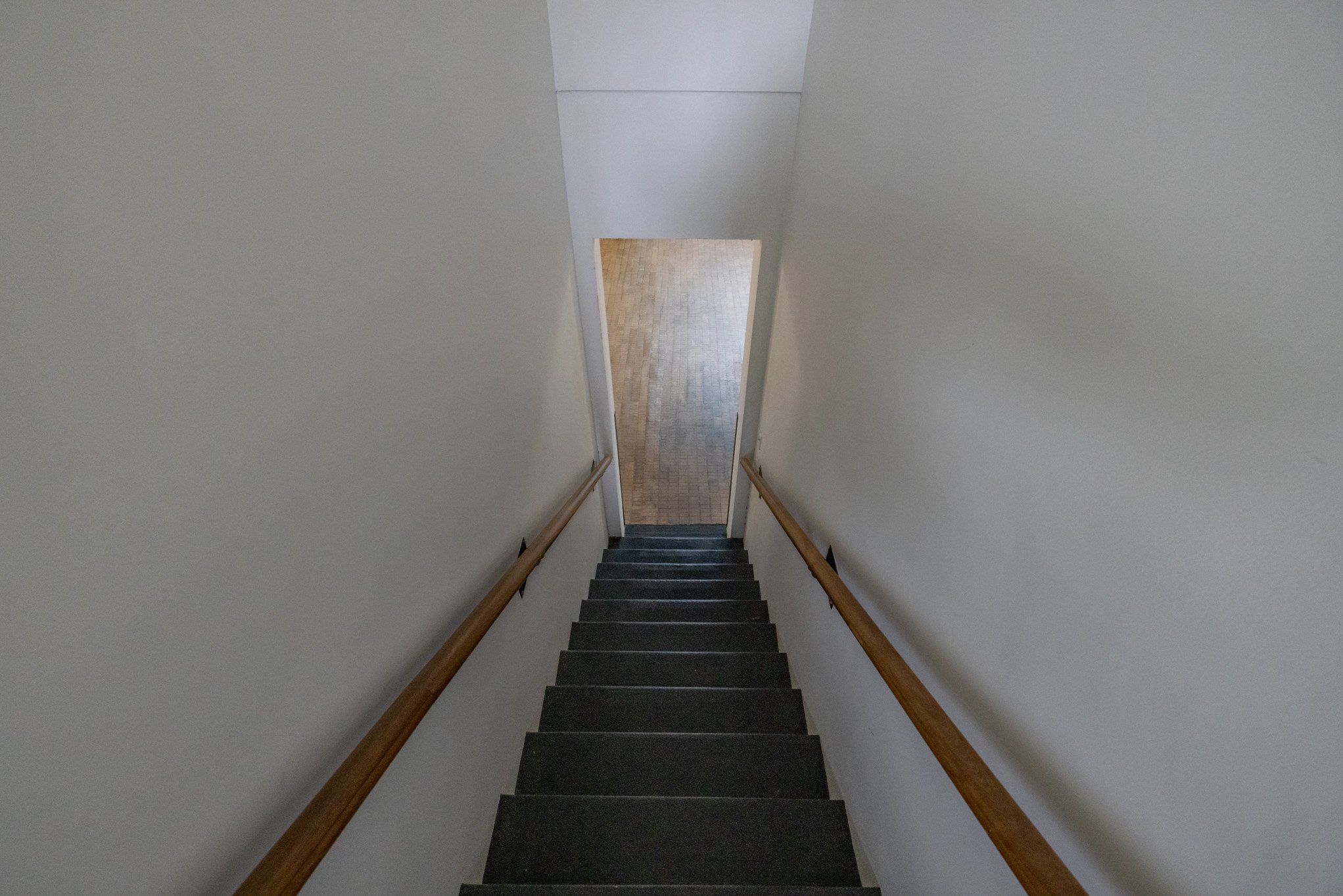 View looking down a staircase with black steps and wooden handrails, leading to a small landing with wood flooring and a door opening into a room with light-colored walls.