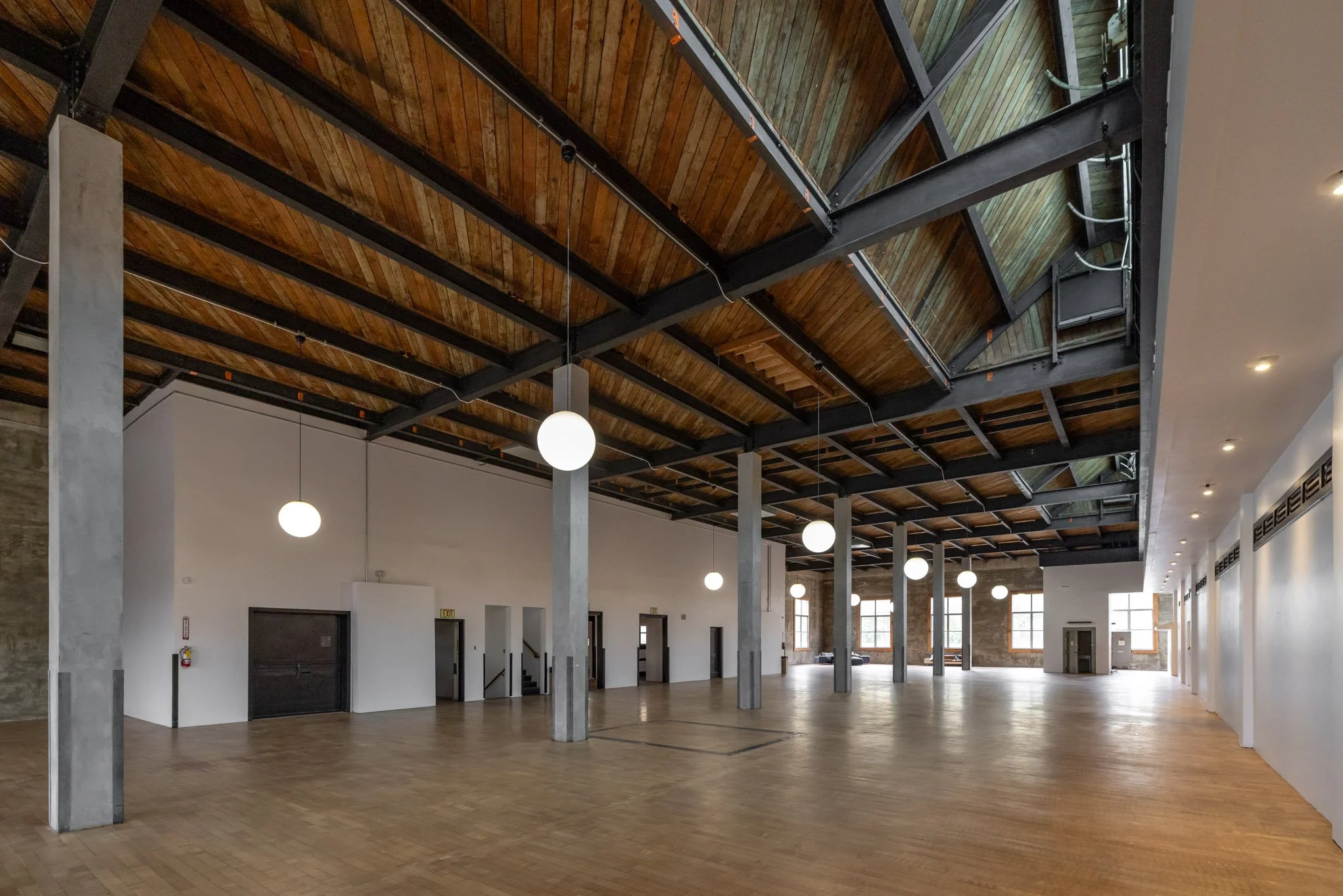 Spacious, empty industrial-style event space with wooden flooring, white walls, large windows, exposed wooden ceiling beams, and hanging spherical lights.