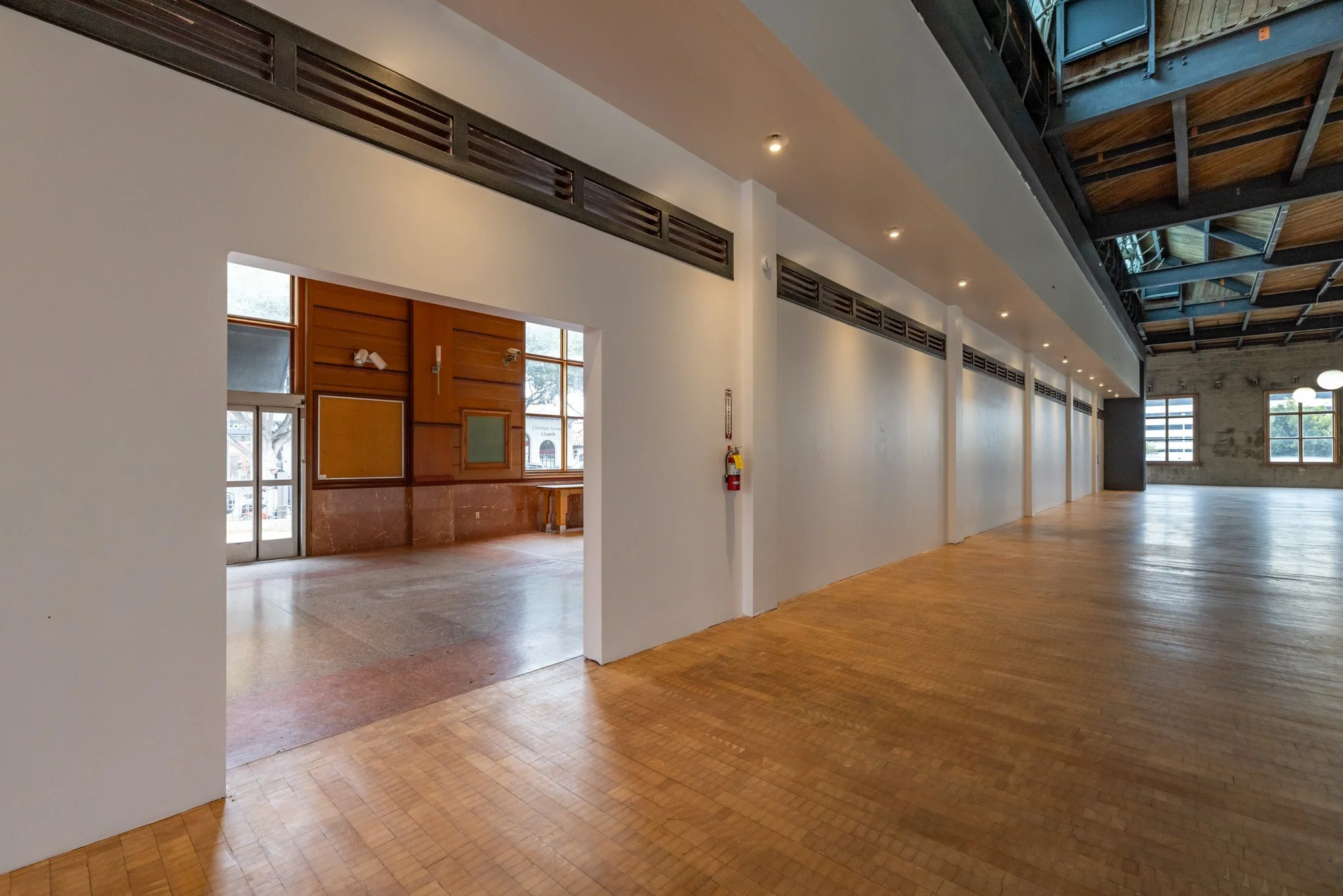 Interior of an empty, spacious room with wooden flooring, white walls, large windows, and a visible fire extinguisher on the wall.