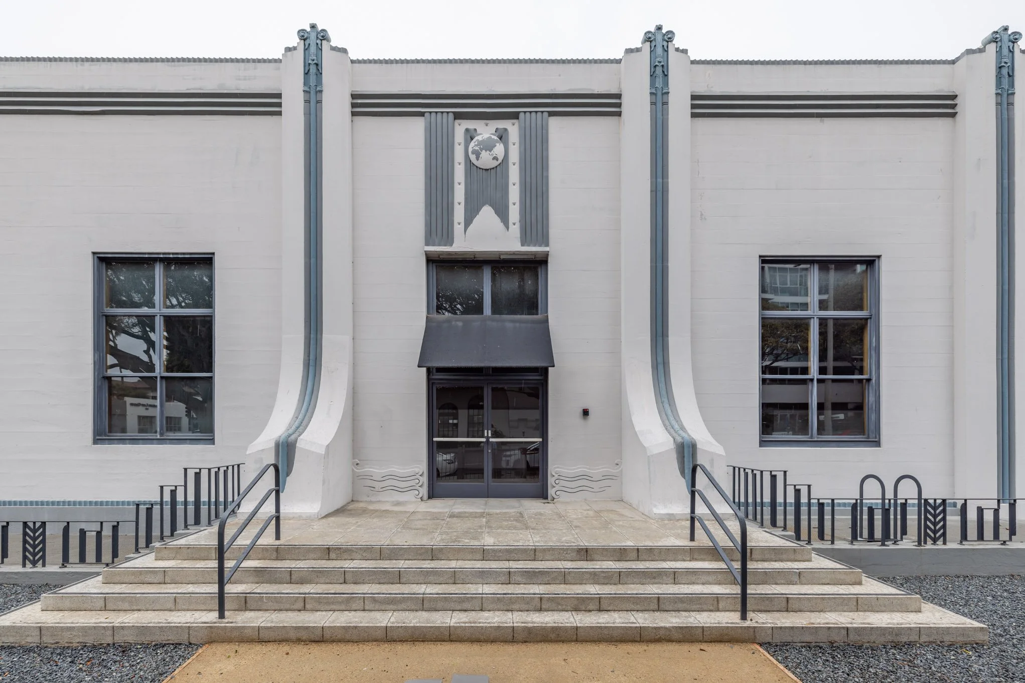 The front of a white Art Deco building with symmetrical design, featuring a globe symbol above the entrance, two large windows, and a staircase leading to glass doors.