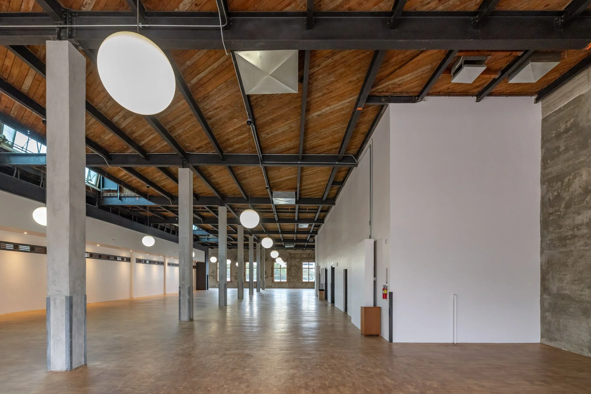 Large, empty industrial-style interior space with wooden ceiling, concrete columns, and hanging spherical lights.