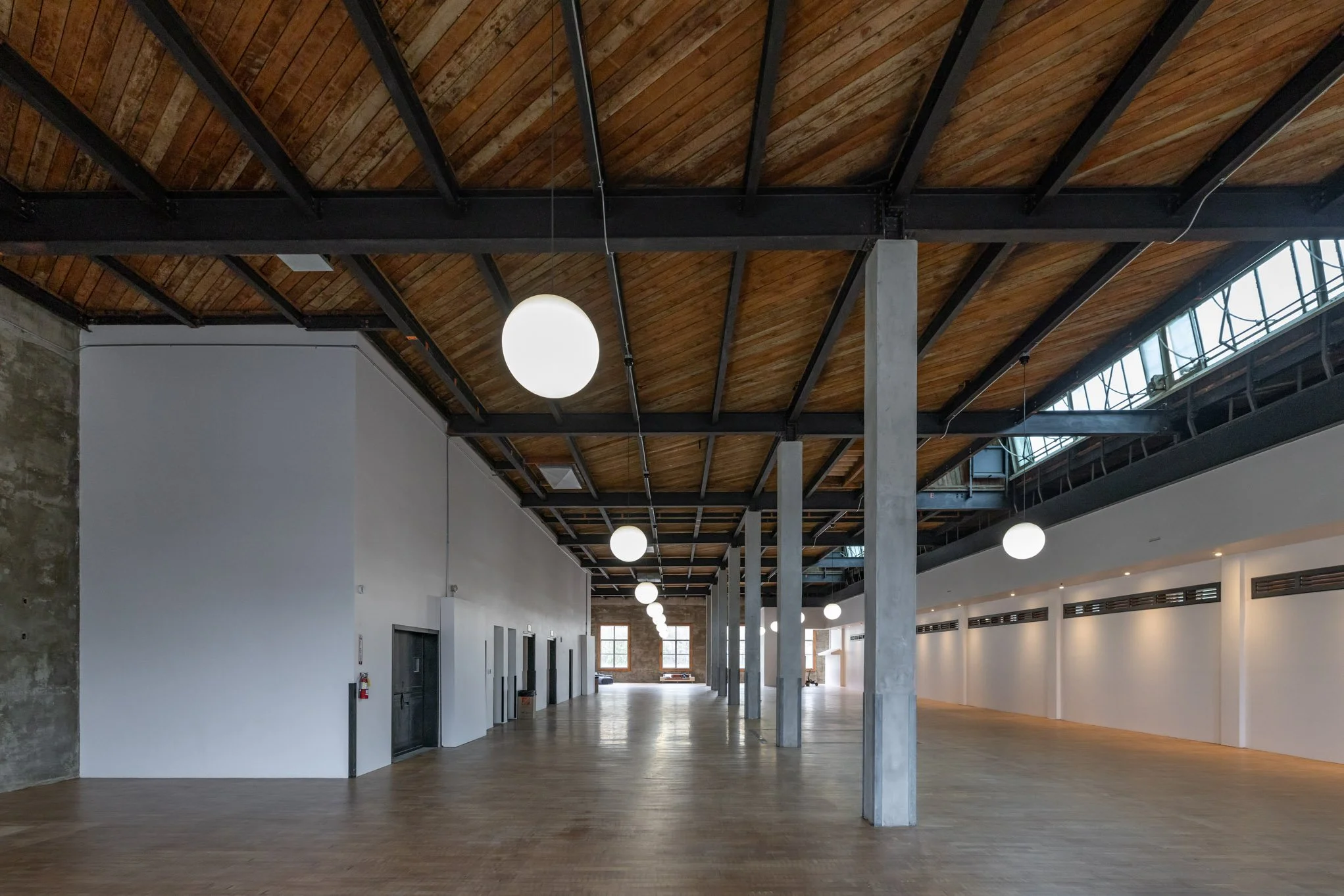 Empty modern interior space with high wooden ceiling, white walls, large windows, and hanging spherical light fixtures.