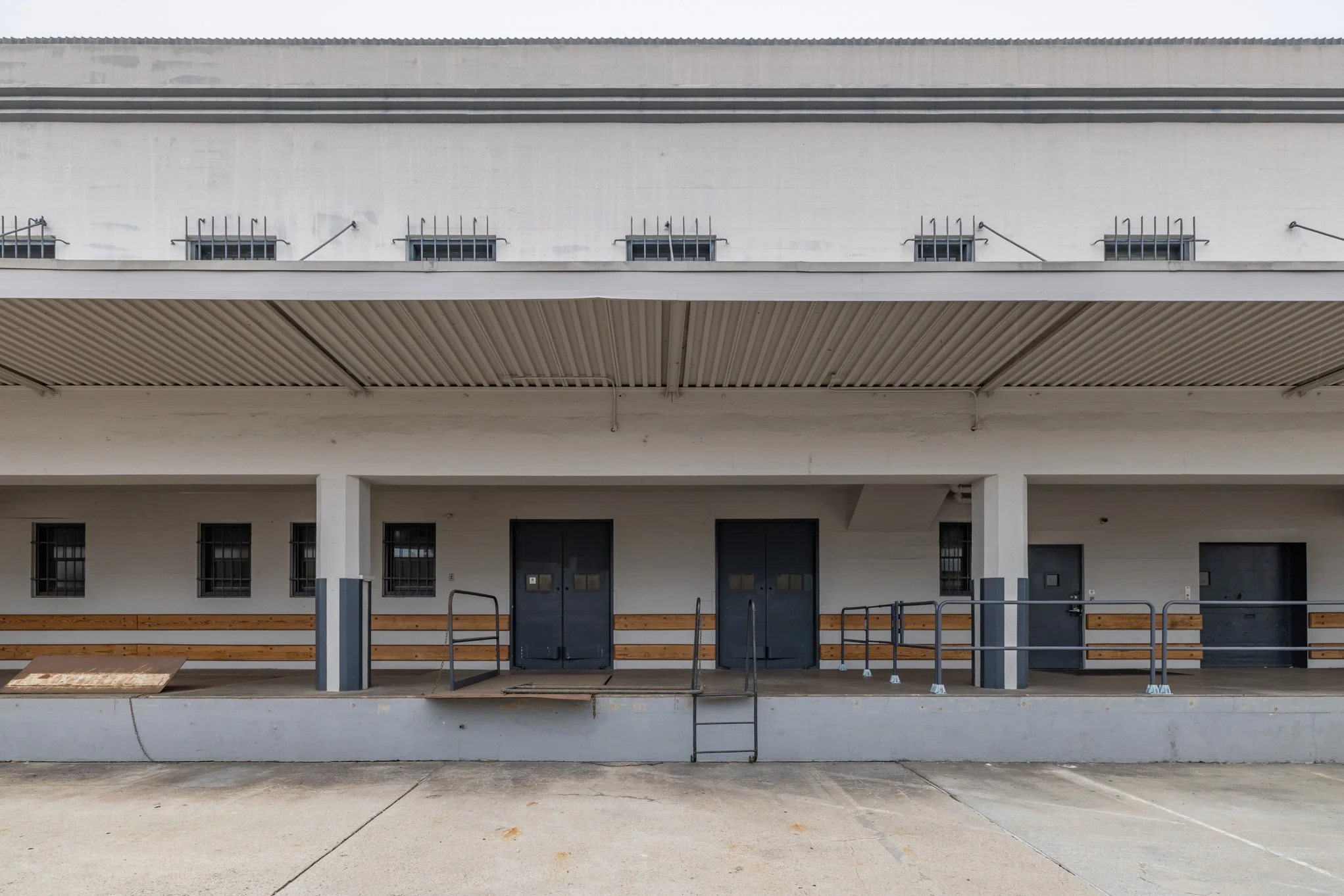 Exterior view of a building with multiple gray doors, windows with bars, and a seating area with wooden benches and metal railings, on a concrete surface.
