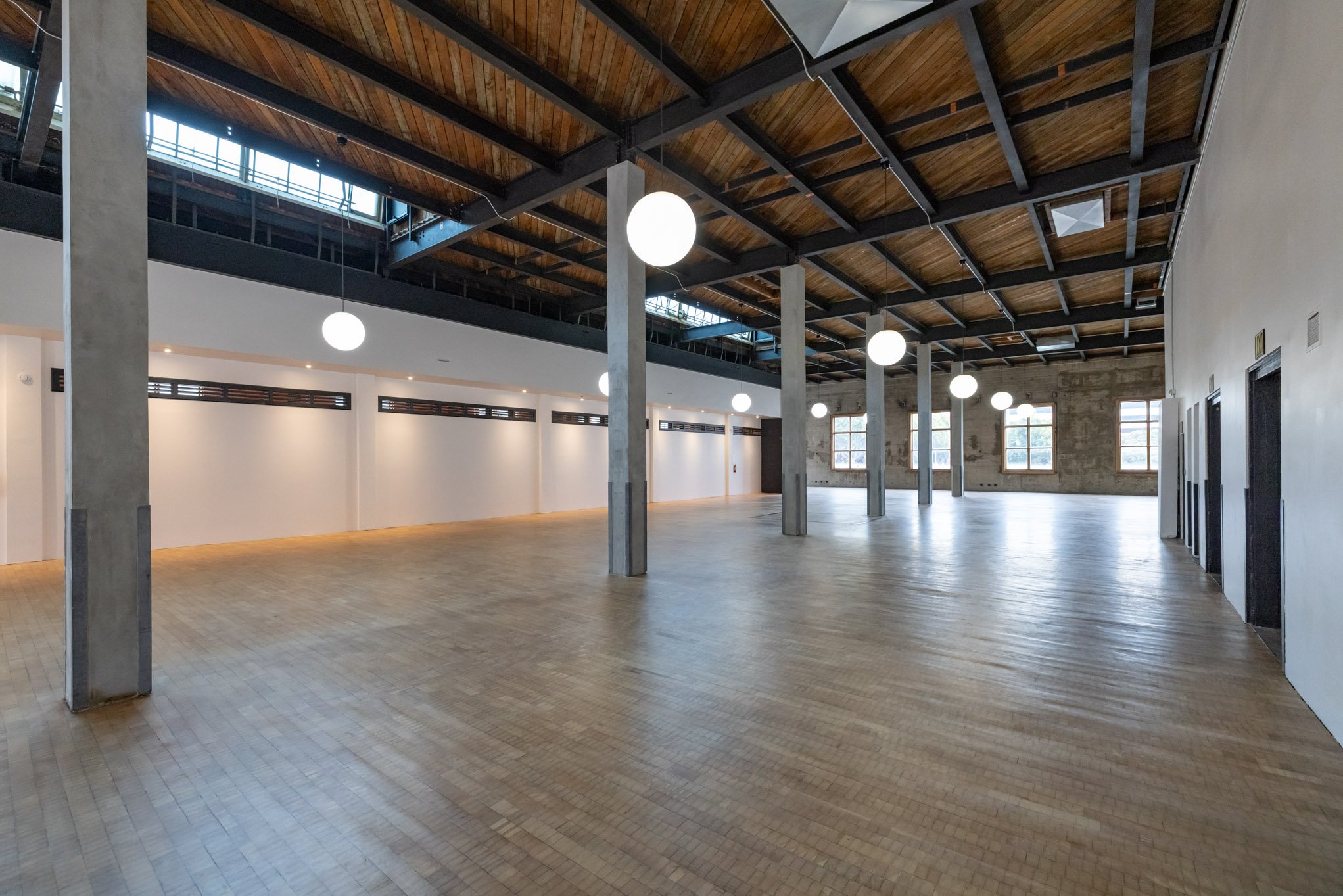 Empty spacious room with wooden floor, white walls, large windows, ceiling with exposed wooden beams, hanging globe lights, and multiple doorways on the right side.