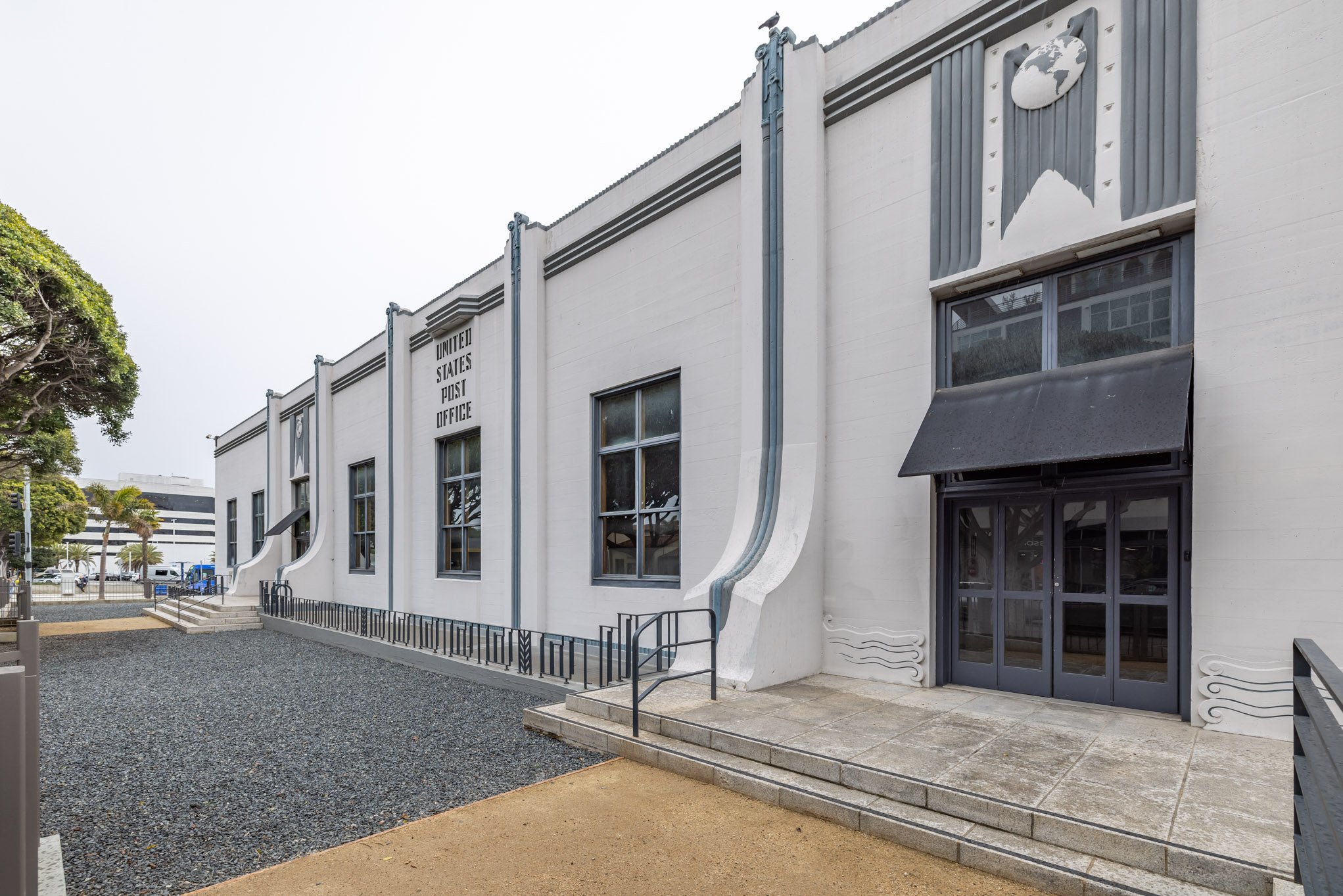 The building is an old United States Post Office with a WWI memorial in an art deco style. The white exterior features stylized decorations, large windows, and a prominent globe emblem at the top. The entrance has steps and a small ramp, with a black