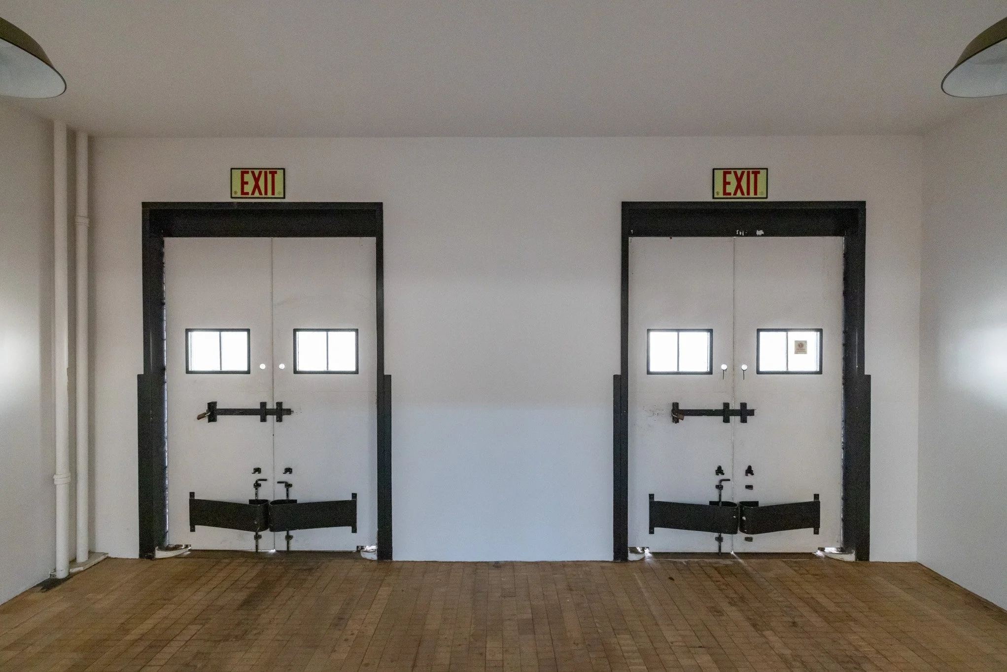Two white exit doors with small windows, black frames, and push bar hardware, in a white-walled room with wood flooring, illuminated by ceiling lights.