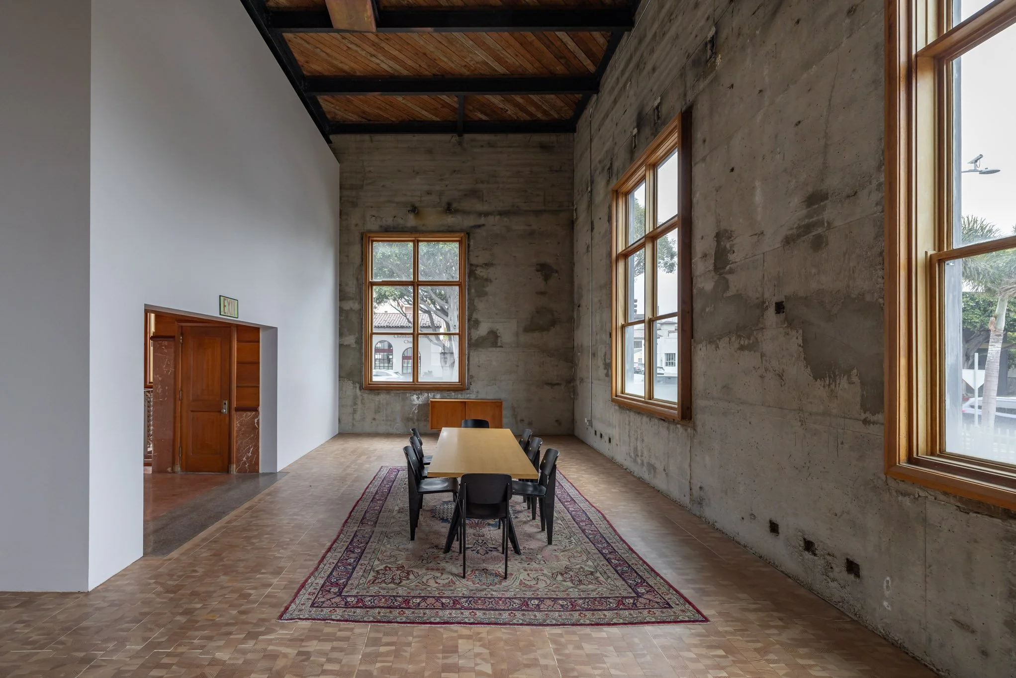A minimalist meeting room with a long wooden table, eight black chairs, a patterned rug, large windows with wooden frames, a concrete wall, and a white wall with a door.
