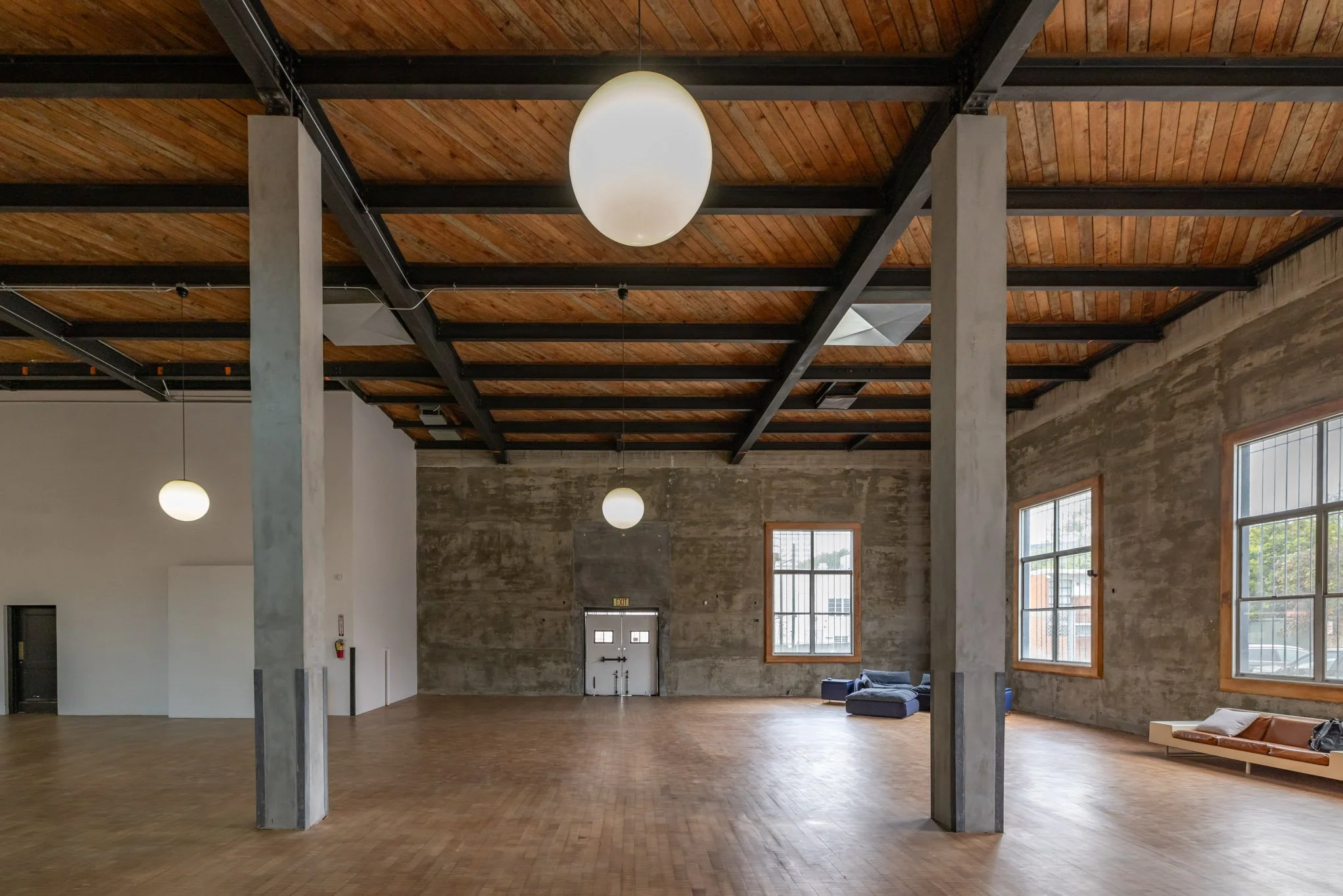 Large empty room with exposed brick walls, wooden ceiling with steel beams, and large windows. There are a few sofas and hanging round lights.