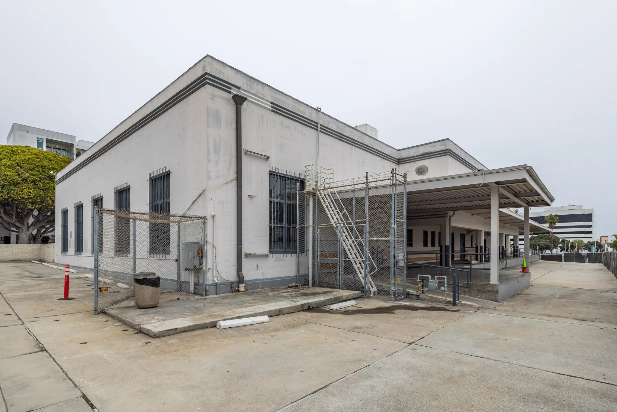 A white building with barred windows, metal stairs, and a fenced off area, with a covered walkway and a parking lot in the foreground, under an overcast sky.