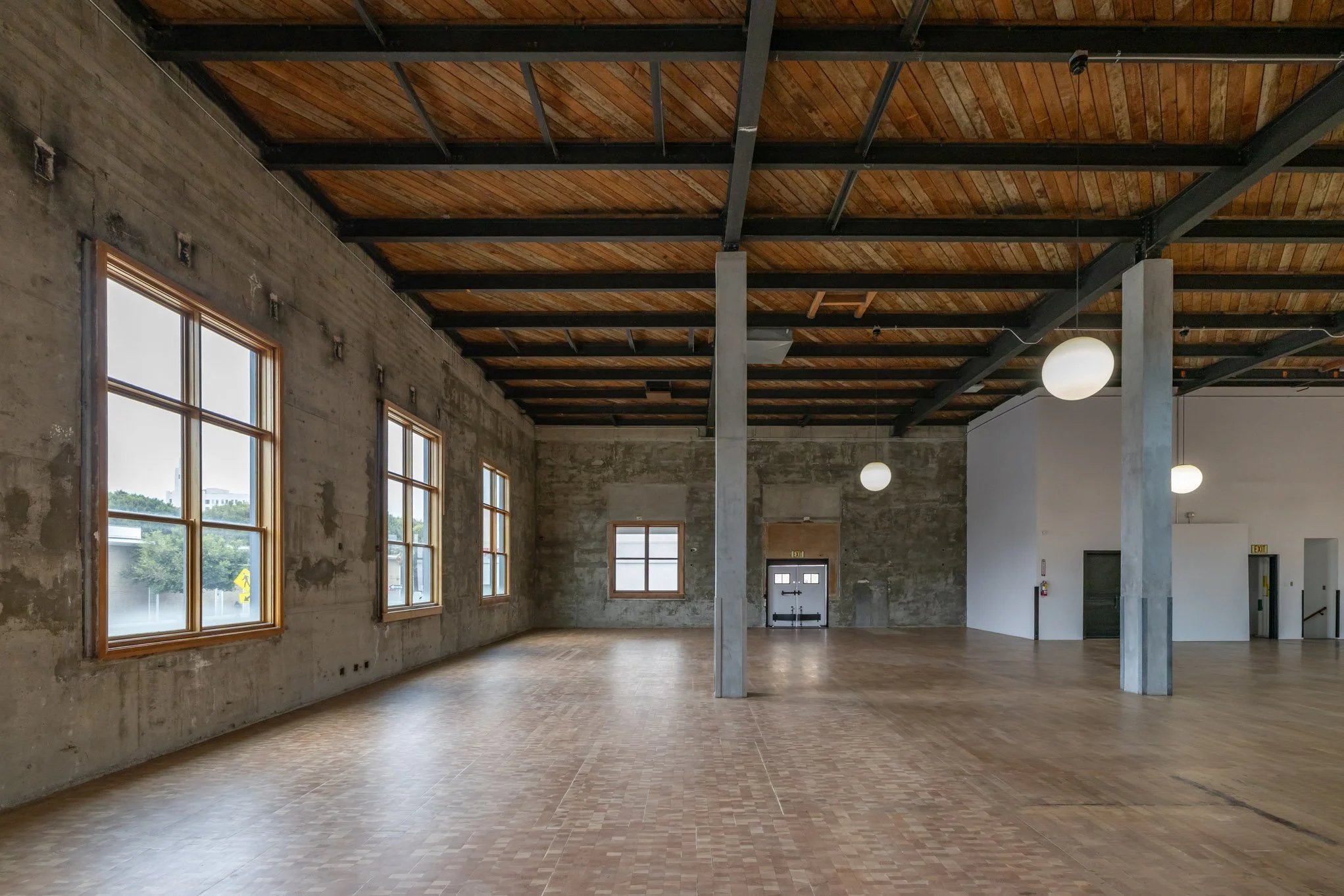 Empty industrial-style room with wooden flooring, concrete walls, large windows, and a wooden ceiling with exposed beams and hanging globe lights.