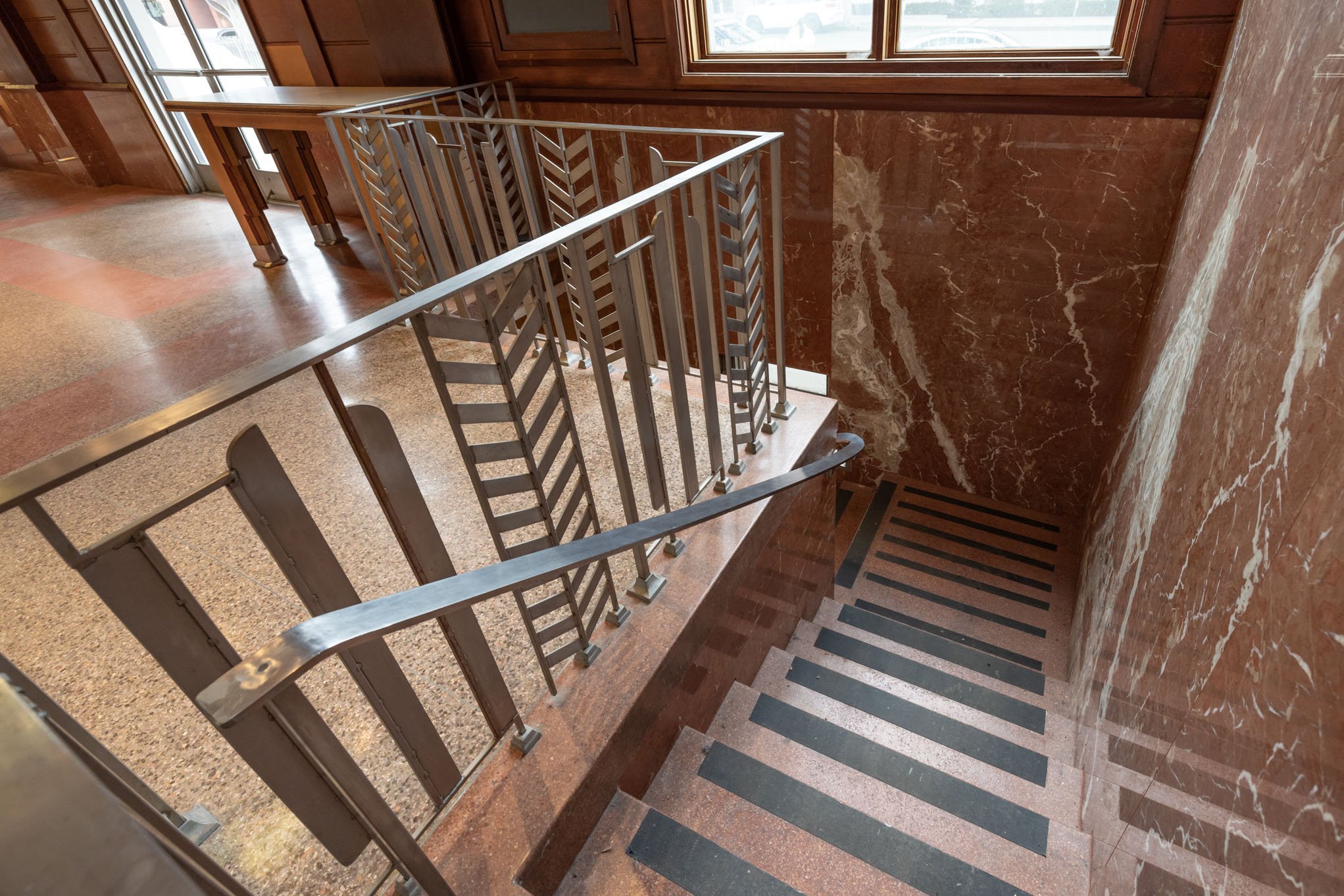 Indoor staircase with metal railing and red marble walls and floor, viewed from the top looking down.