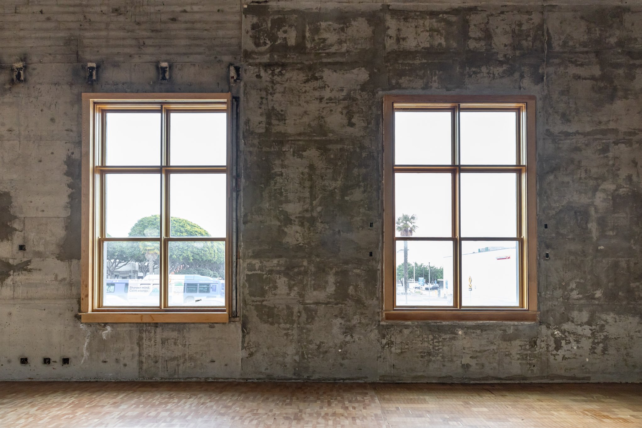 Two large, square, wooden-framed windows in an unfinished concrete wall with a view of trees, cars, and a street outside.